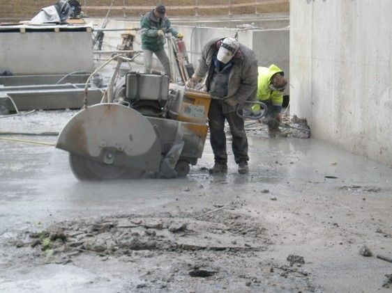 Drei Bauarbeiter benutzen eine handgeführte Betonsäge, um einen nassen, mit Schlamm bedeckten Betonboden zu durchtrennen.