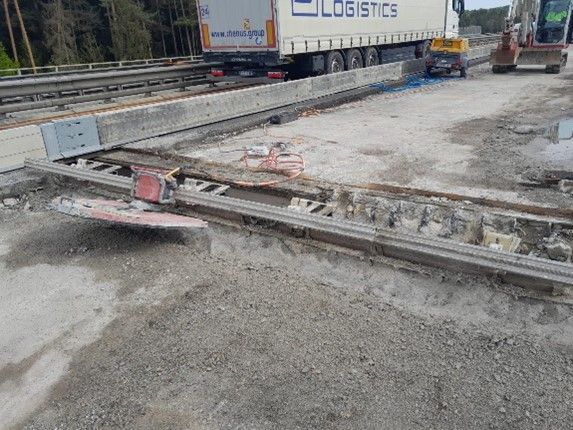 Eine Baustelle, auf der eine Betonbrückenfuge mit einer Metallplatte repariert wird und schwere Baumaschinen zu sehen sind.