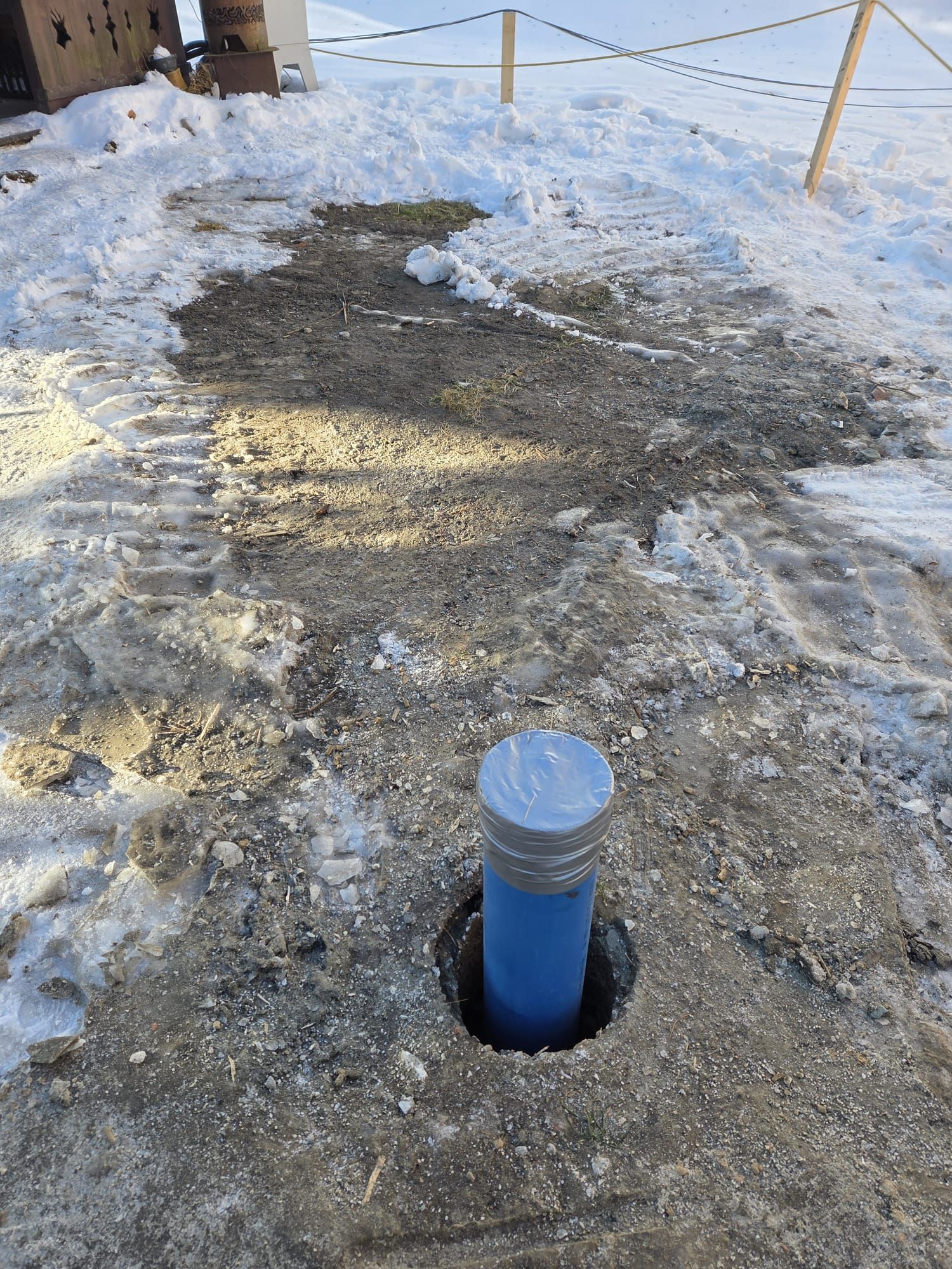 Aus einem Loch in einem schneebedeckten, kiesigen Stück Boden ragt ein blaues Rohr mit einer Metallkappe.