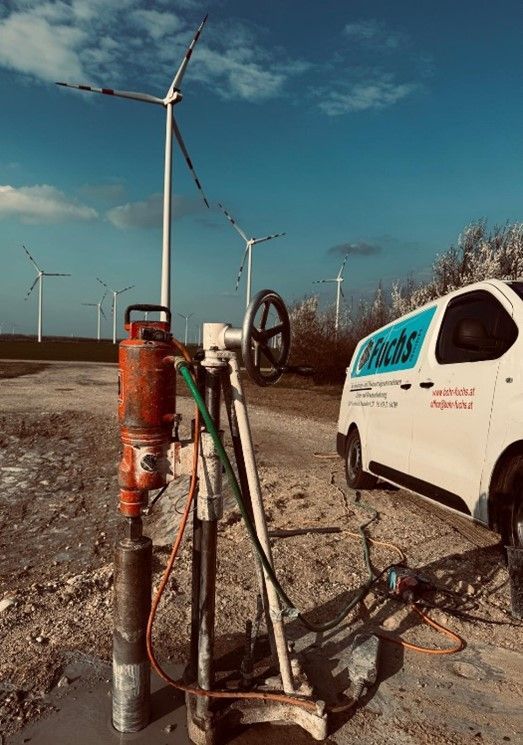 Auf Kies steht eine Kernbohranlage neben einem weißen Lieferwagen; im strahlend blauen Himmel sind Windkraftanlagen zu sehen.