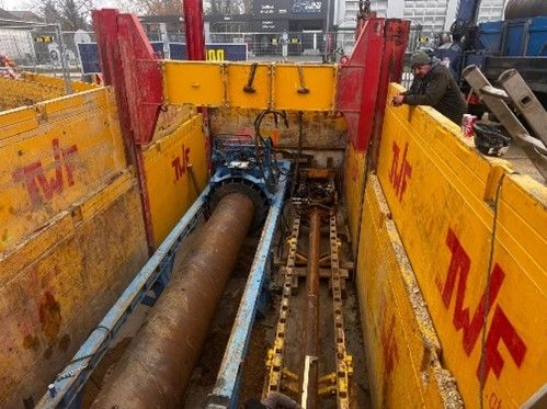 Ein Bauarbeiter überwacht eine Horizontalbohrmaschine in einem tiefen, mit gelben Stützplatten ausgekleideten Graben.