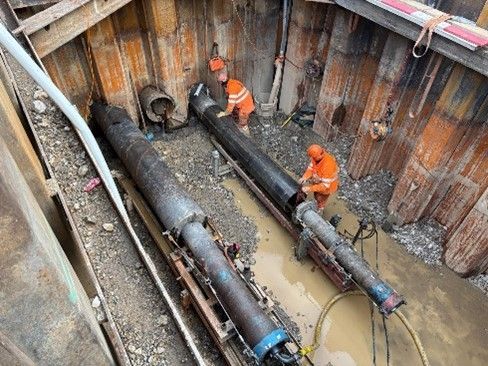 Zwei Arbeiter in orangefarbener Warnkleidung arbeiten an großen horizontalen Rohren in einem tiefen Baugraben.