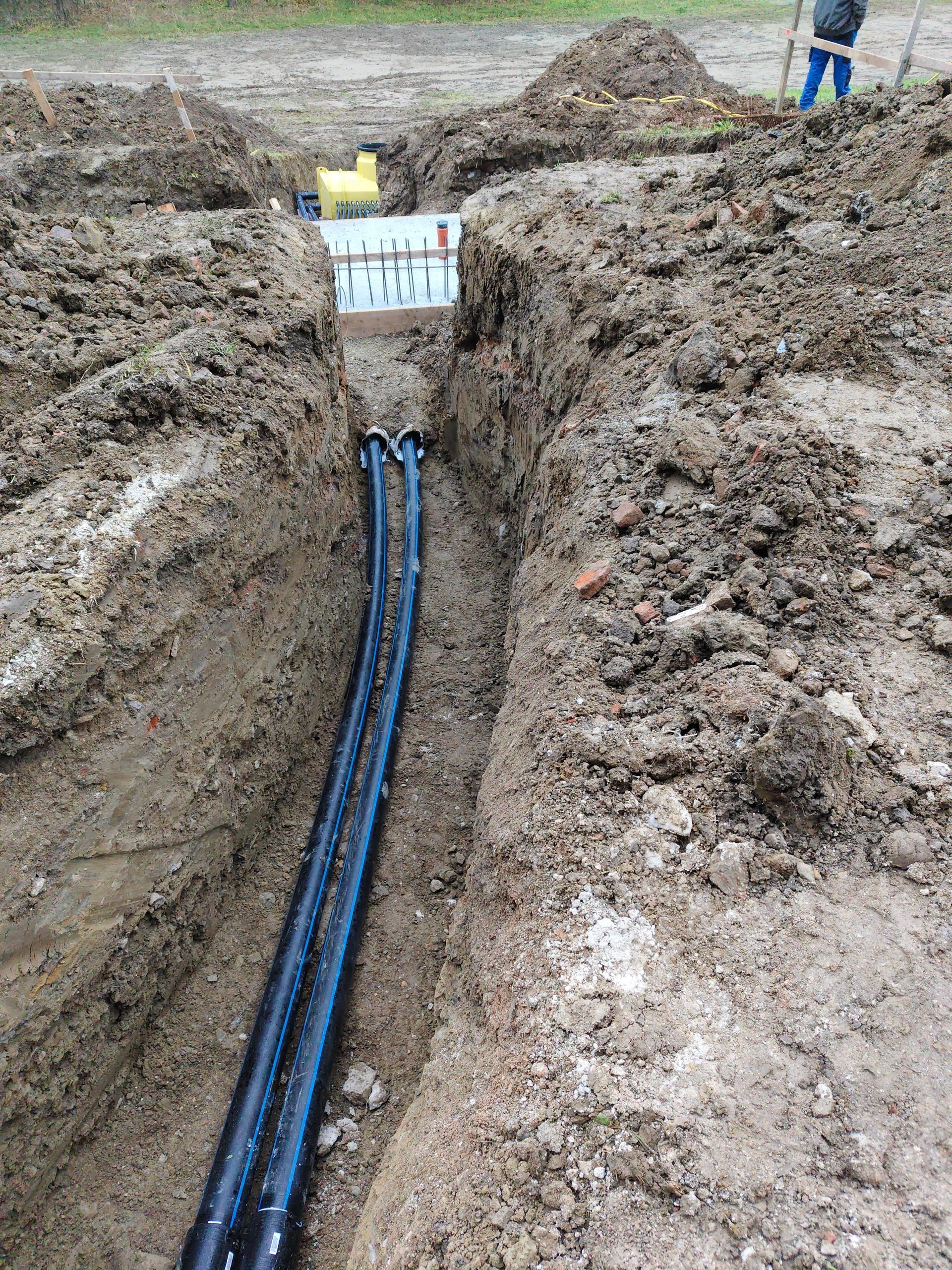 Am Grund eines tiefen Erdgrabens, der zu einem weißen Bauwerk führt, liegen zwei schwarze Rohre, über dem eine Person steht.