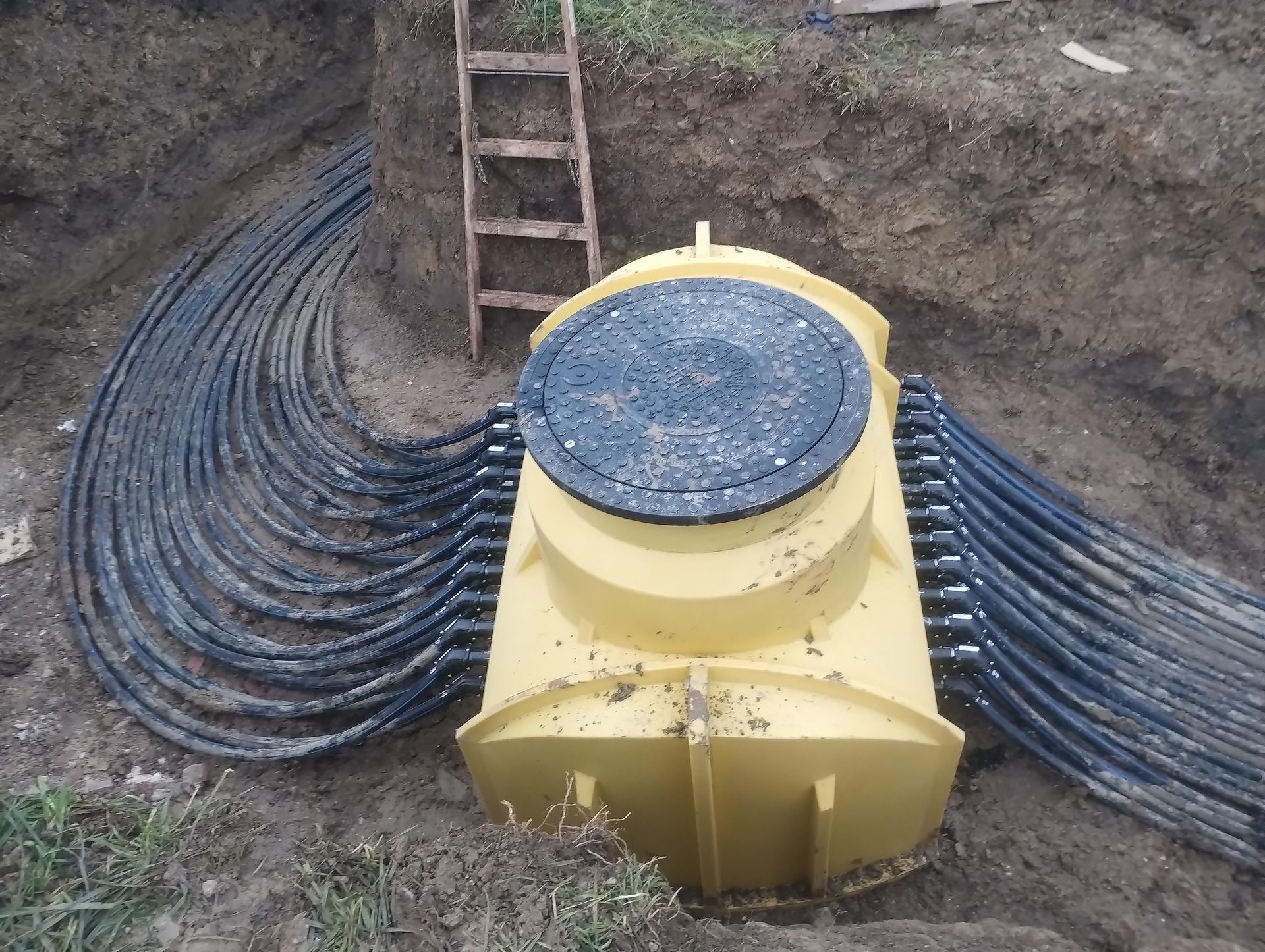 Ein gelber unterirdischer Verteilerkasten ist mit mehreren schwarzen Leitungsrohren in einem Graben verbunden