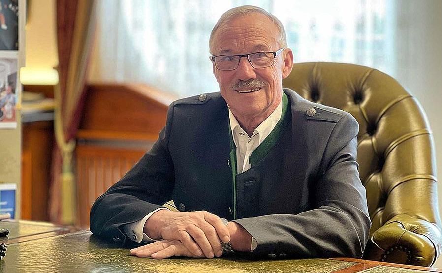 Eine lächelnde Person in Jackett und Weste sitzt mit verschränkten Händen an einem Schreibtisch in einem Büro.