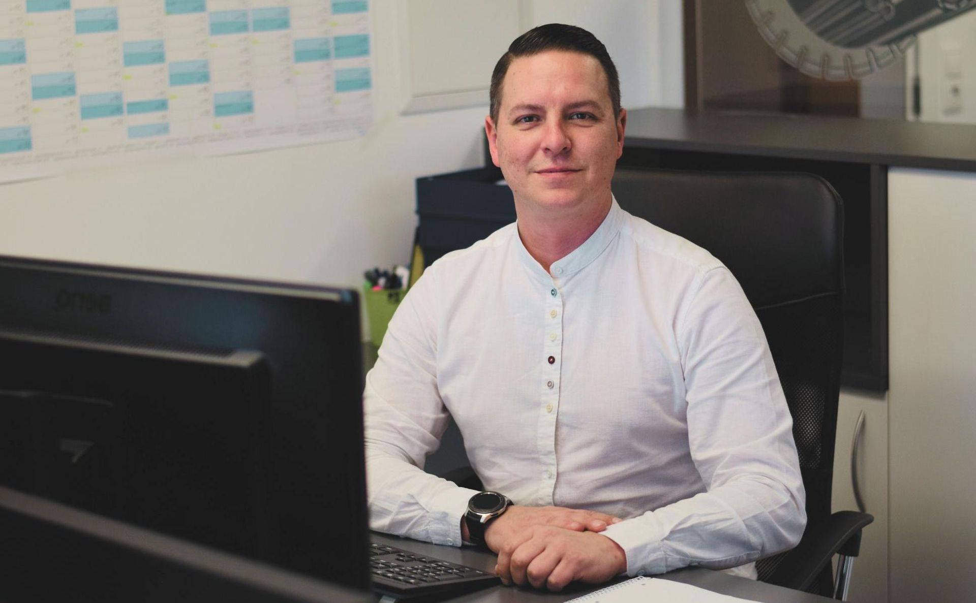 Ein Geschäftsmann sitzt in einem Büro an einem Schreibtisch vor einem Computer und trägt ein weißes Hemd mit Knöpfen.