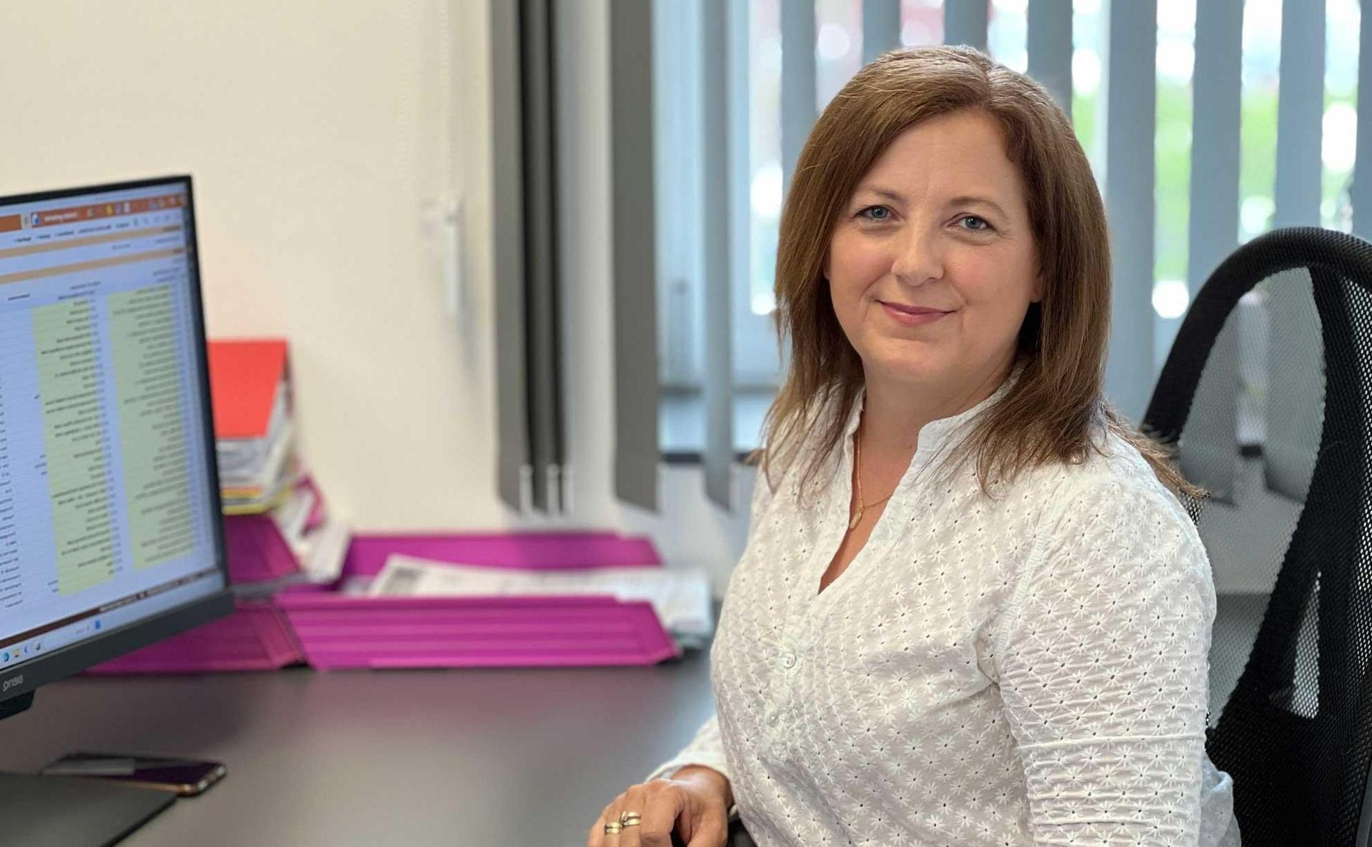 Eine lächelnde Person sitzt an einem Schreibtisch vor einem Computermonitor und trägt ein weißes Oberteil mit Spitzenmuster.