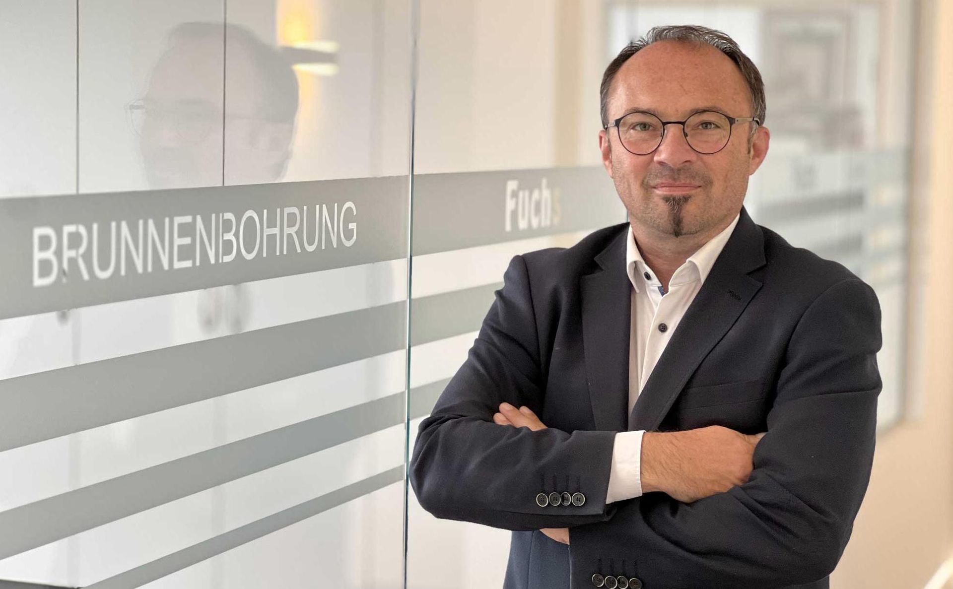 Ein Profi steht mit verschränkten Armen vor einer Glaswand mit der Aufschrift „BRUNNENBOHRUNG“.