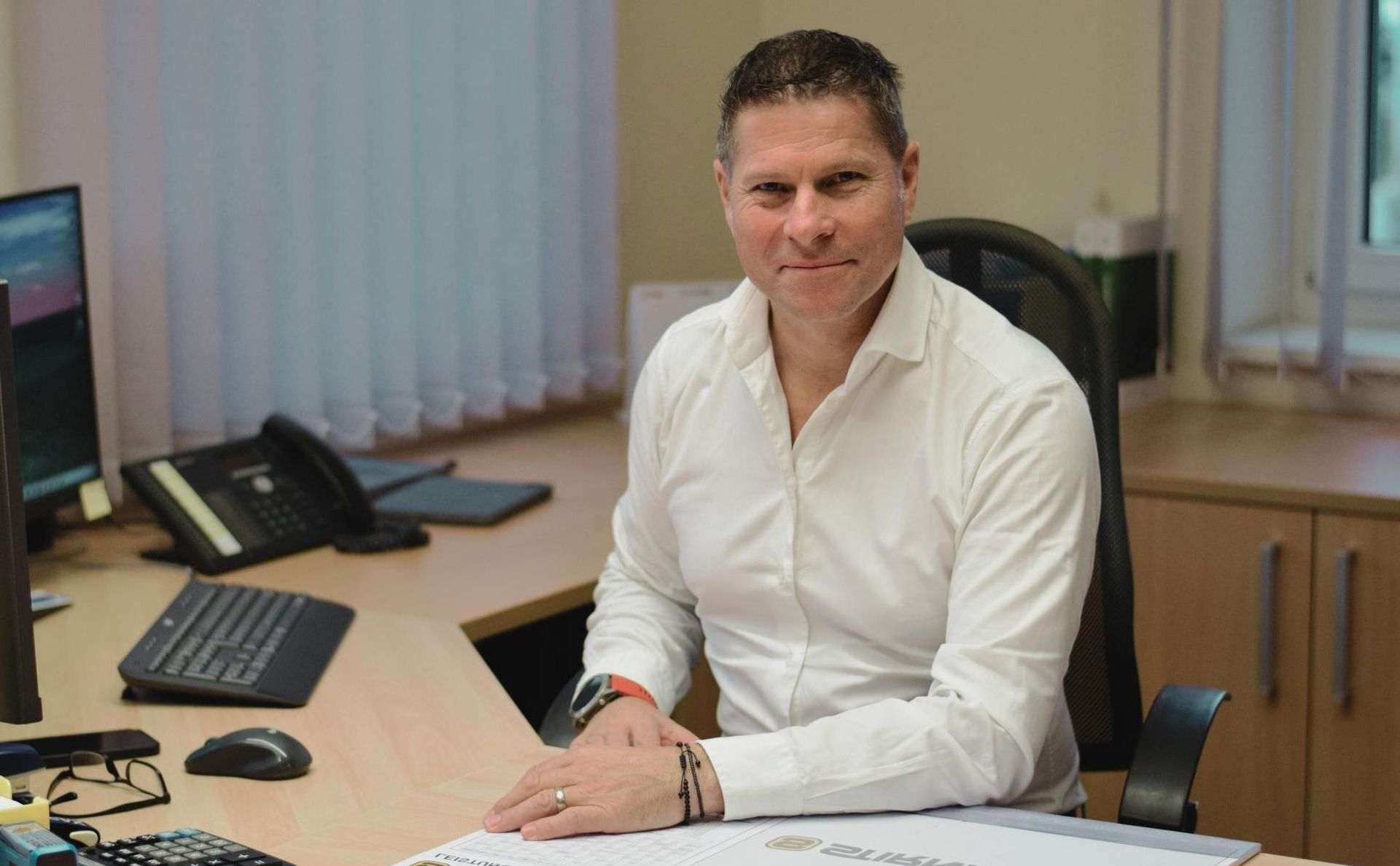 Ein Mann in einem weißen Hemd mit Knöpfen sitzt an einem Schreibtisch und blickt mit einem leichten Lächeln in die Kamera.