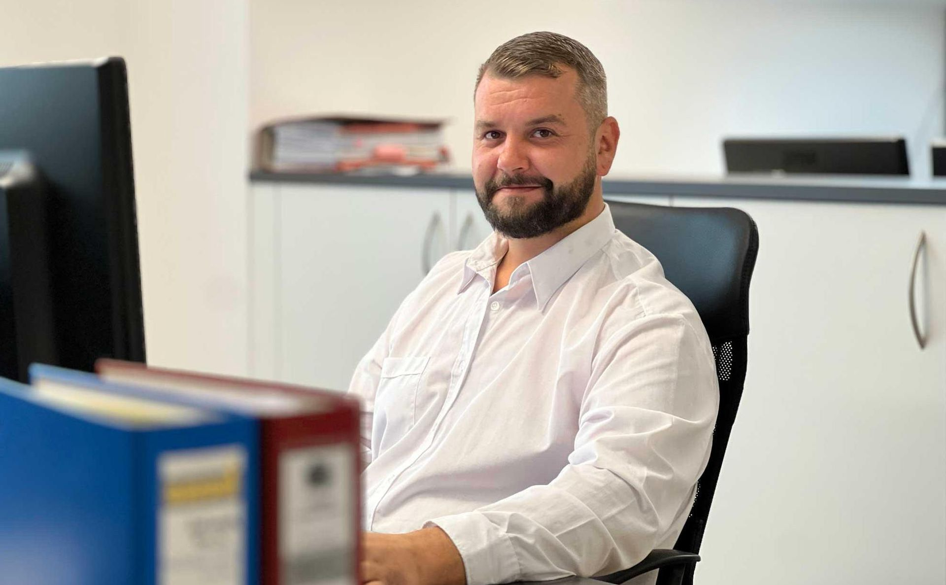 Eine Person mit Bart, die ein weißes Hemd mit Knöpfen trägt, sitzt an einem Schreibtisch im Büro