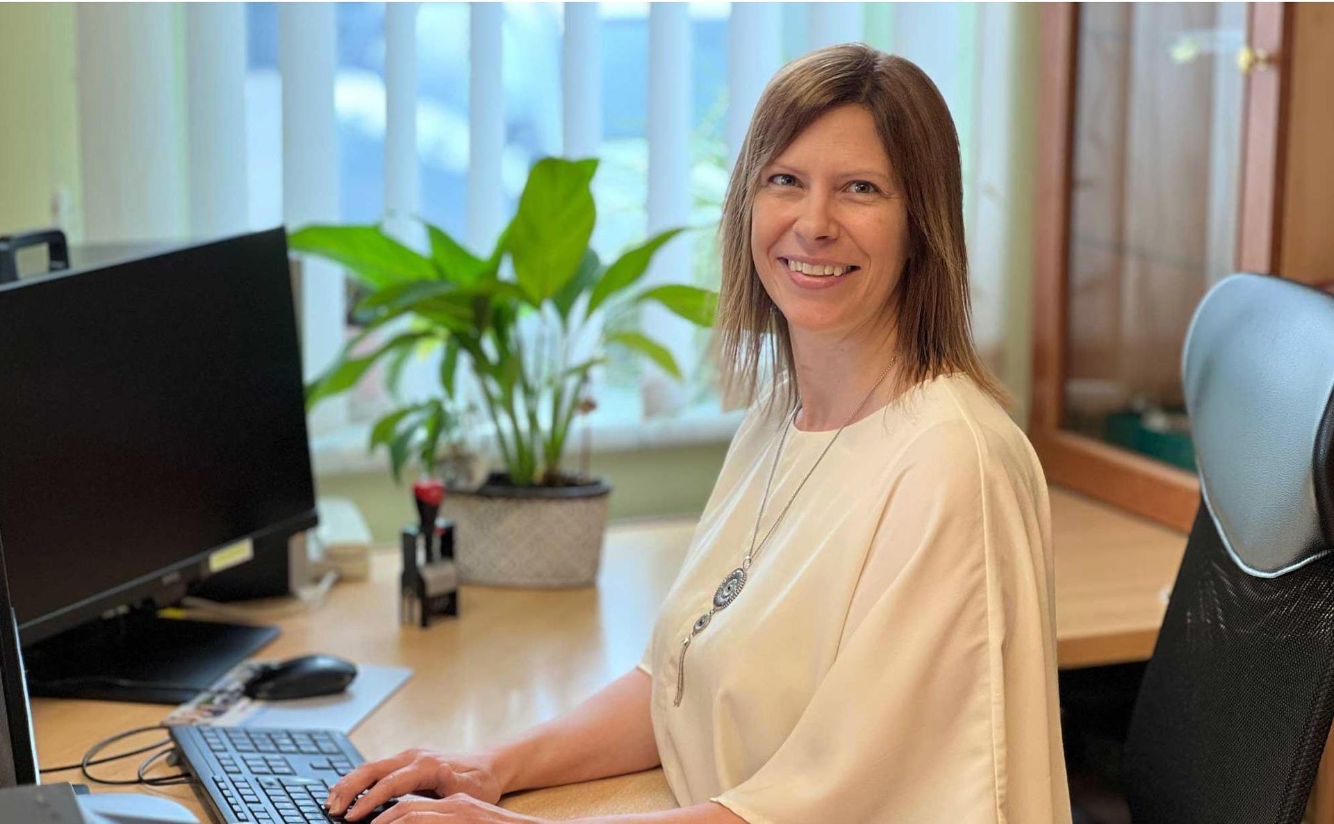 Eine berufstätige Frau lächelt an ihrem Schreibtisch neben einem Computer und einer Topfpflanze im Büro.