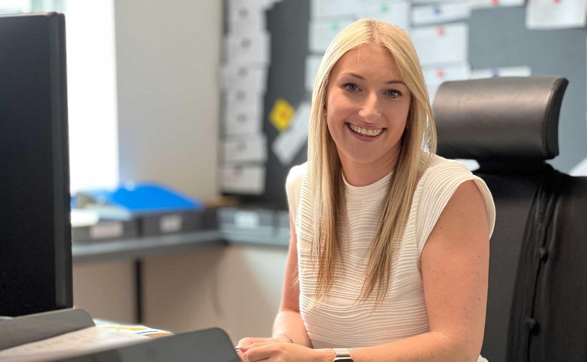 Eine lächelnde Person mit langen blonden Haaren sitzt an einem Schreibtisch in einem Büro 