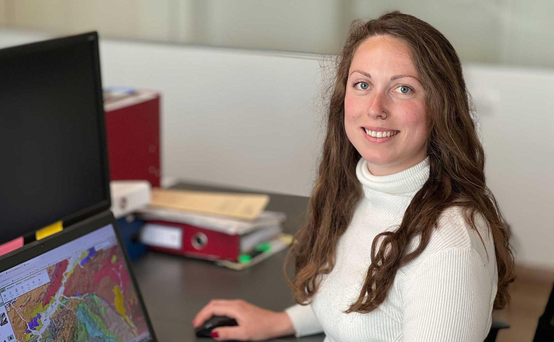 Eine Person mit langen braunen Haaren und einem weißen Rollkragenpullover sitzt an einem Schreibtisch 