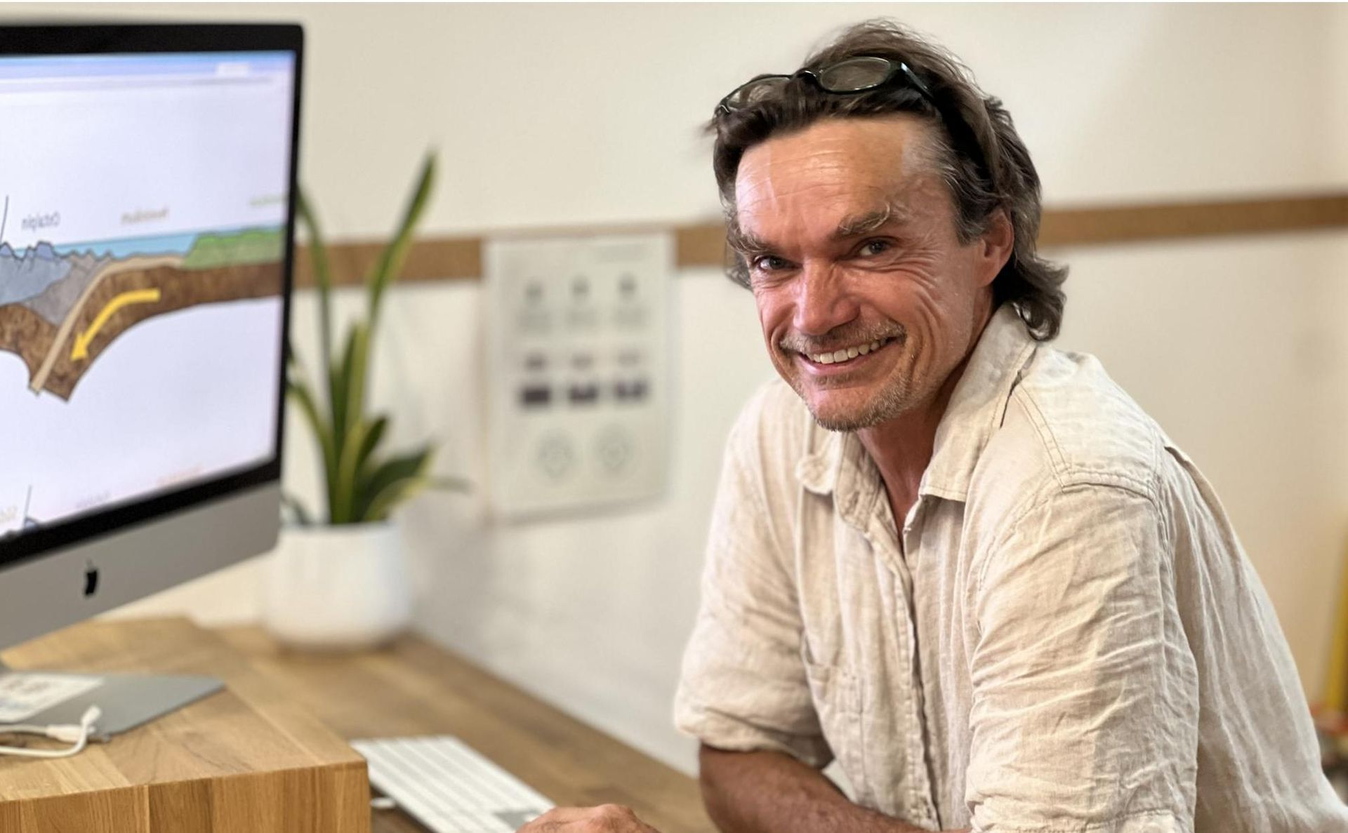 Eine Person lächelt an einem Schreibtisch vor einem Computerbildschirm, auf dem ein geologisches Diagramm zu sehen ist.