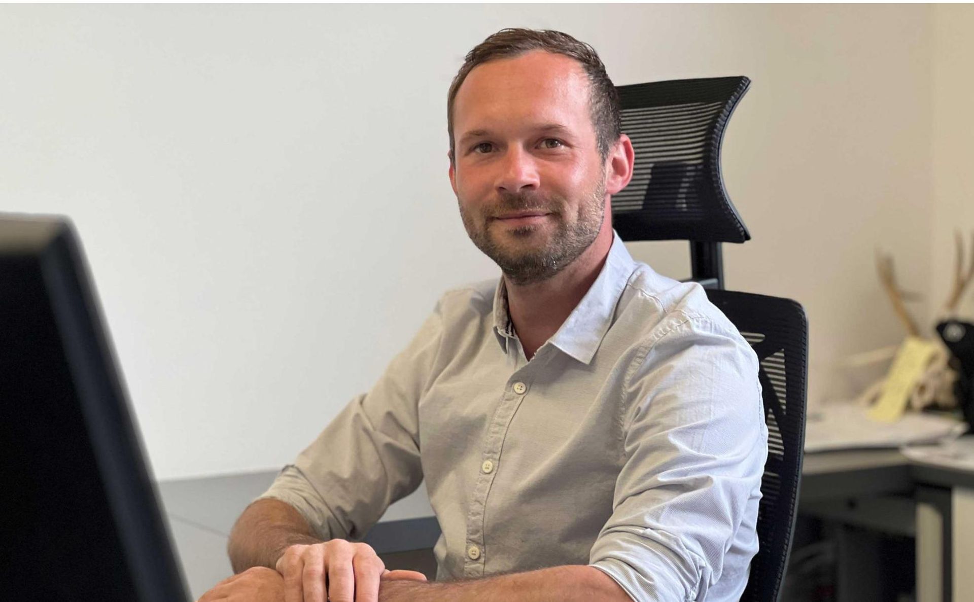 Eine Person in einem hellen Hemd mit Knöpfen sitzt in einem Büro an einem Schreibtisch mit Computermonitor.