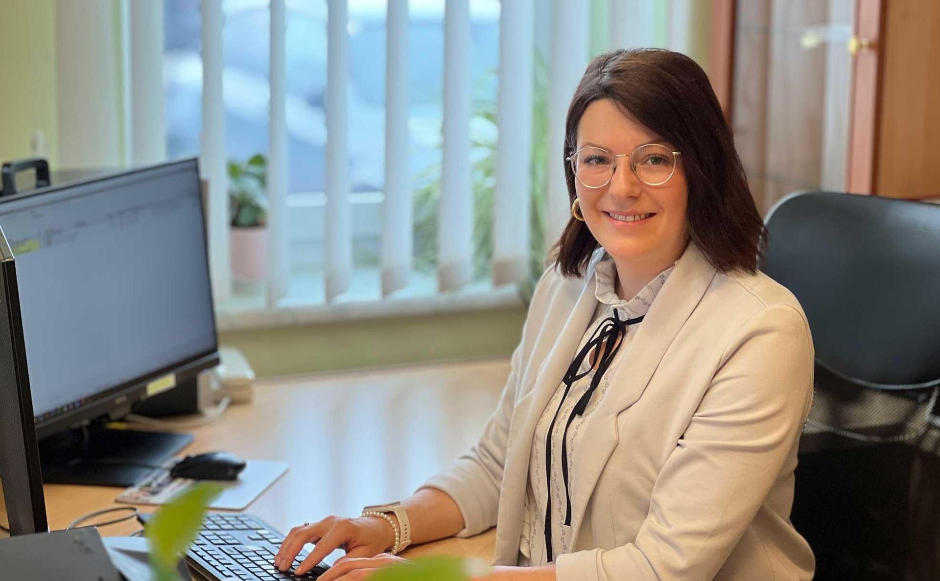 Eine Person mit Brille und hellem Blazer sitzt an einem Schreibtisch und tippt in einem Büro auf einer Tastatur.