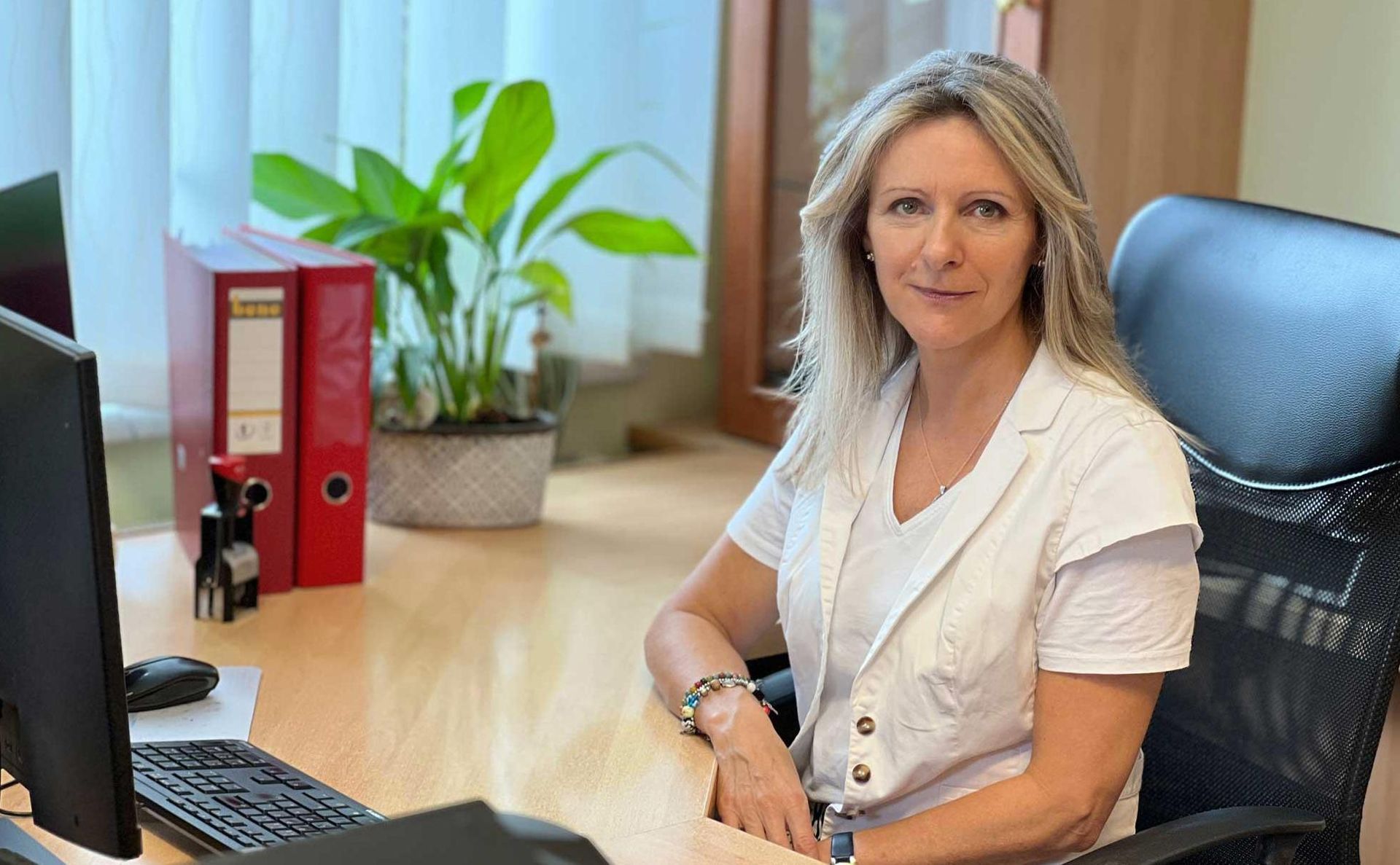 Eine berufstätige Frau in einer weißen Bluse sitzt an einem Schreibtisch neben einem Computer