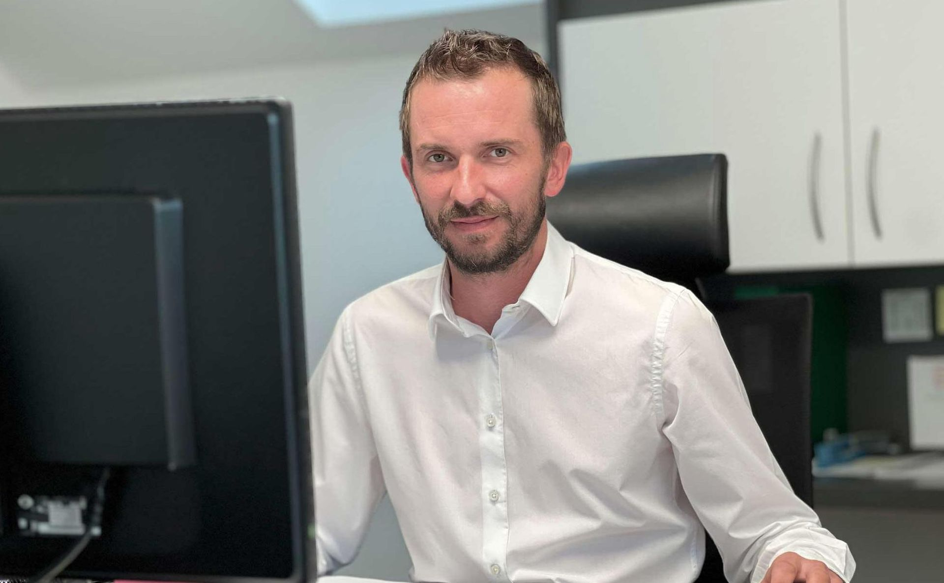 Eine Person in einem weißen Hemd mit Knöpfen sitzt an einem Schreibtisch vor einem Computermonitor und blickt in die Kamera.