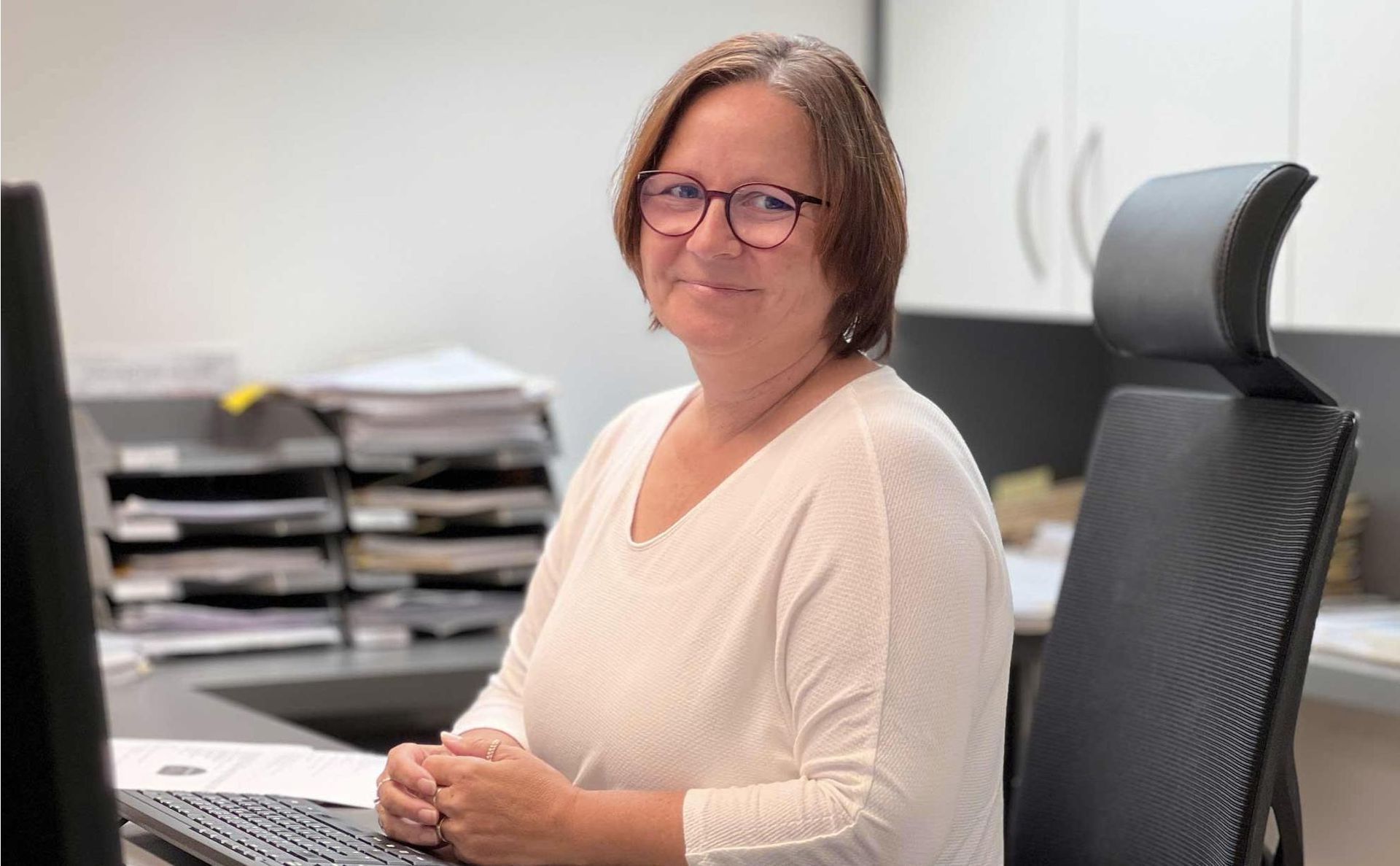 Eine Person mit Brille und einem hellen, langärmeligen Hemd sitzt an einem Schreibtisch; 