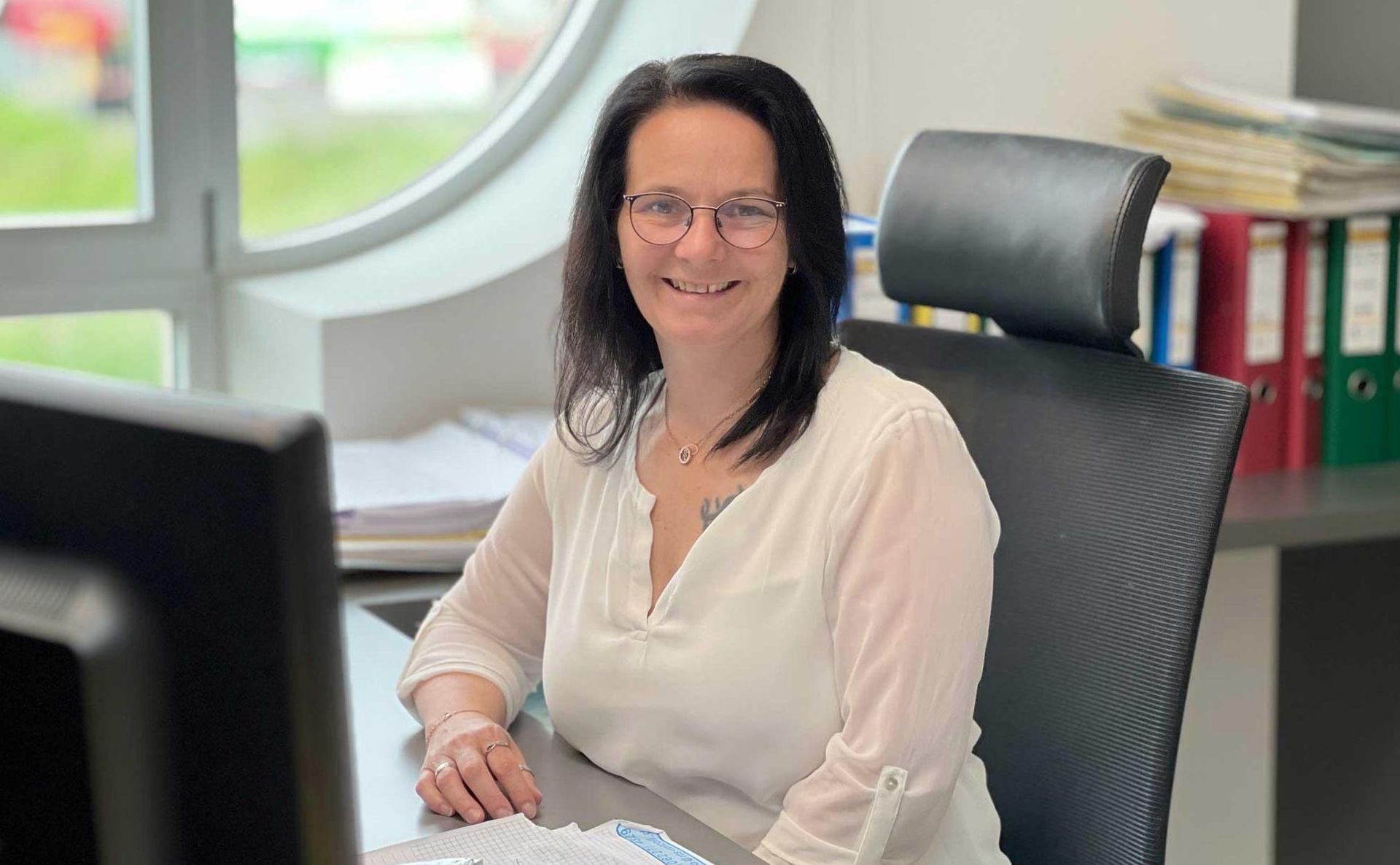 Eine Person mit Brille und weißer Bluse sitzt an einem Schreibtisch; neben ihr stehen Ordner und ein Computermonitor.