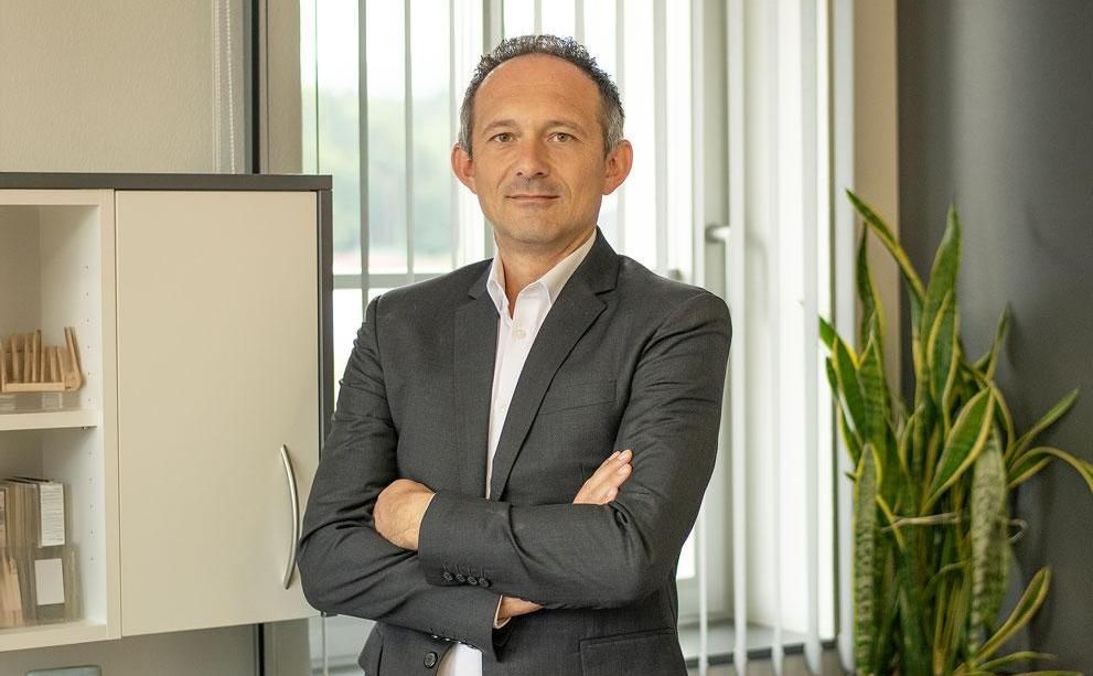 Eine Person in einem grauen Blazer und einem weißen Hemd steht mit verschränkten Armen in einem Büro 