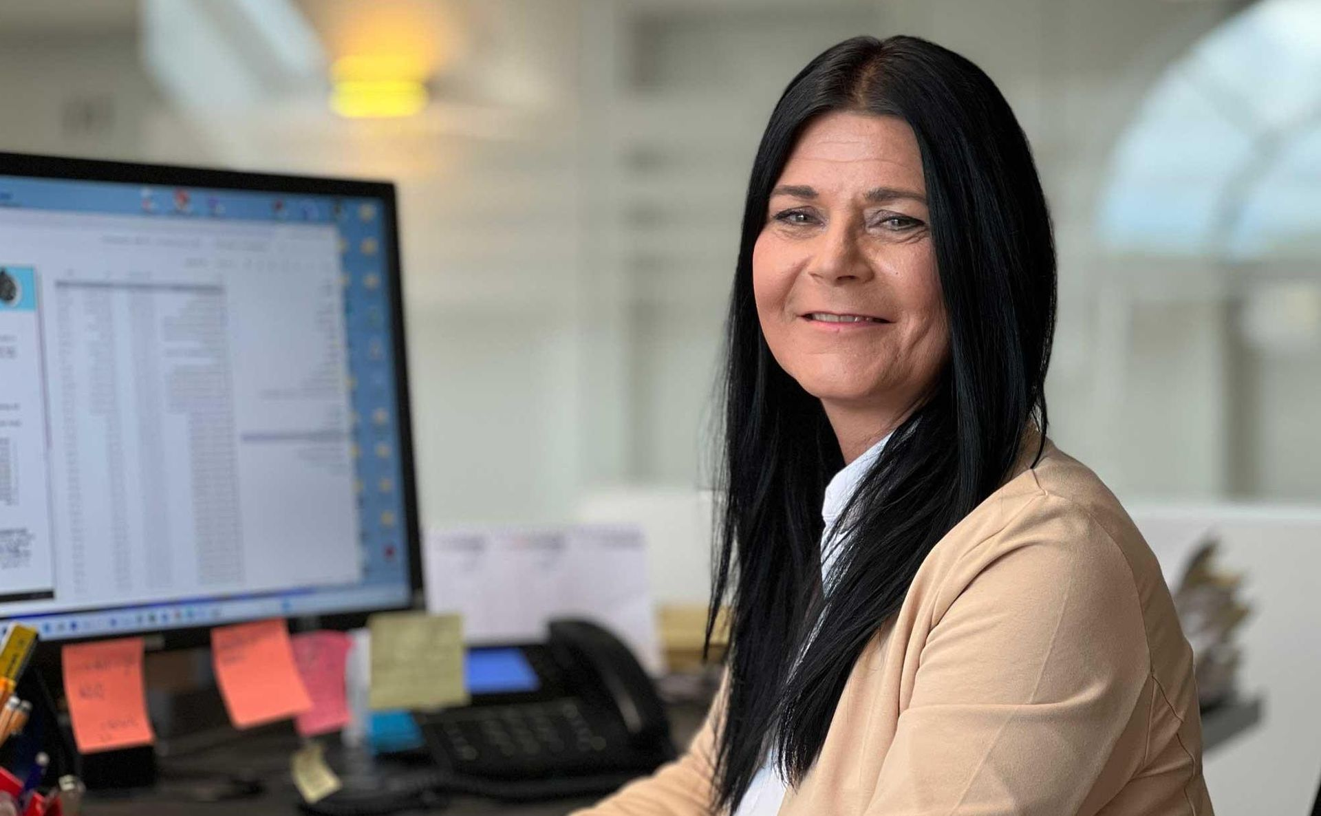 Eine lächelnde Person in einer beigen Strickjacke sitzt an einem Schreibtisch mit Computermonitor