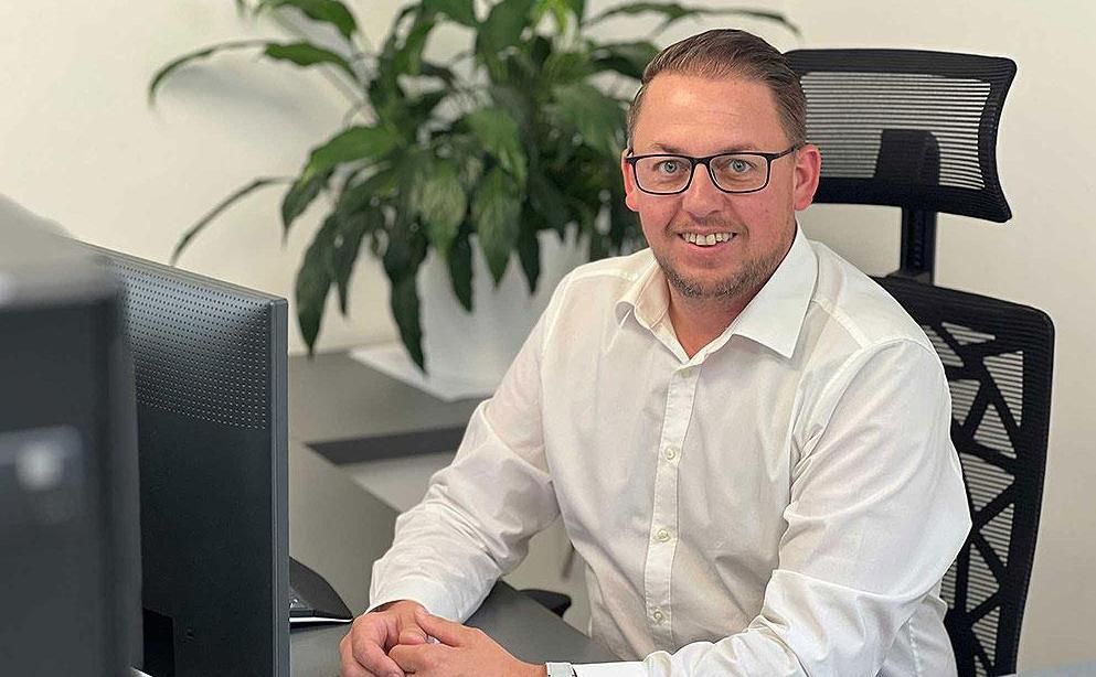 Eine Person in einem weißen Hemd mit Knöpfen sitzt lächelnd an einem Schreibtisch mit einem Computer