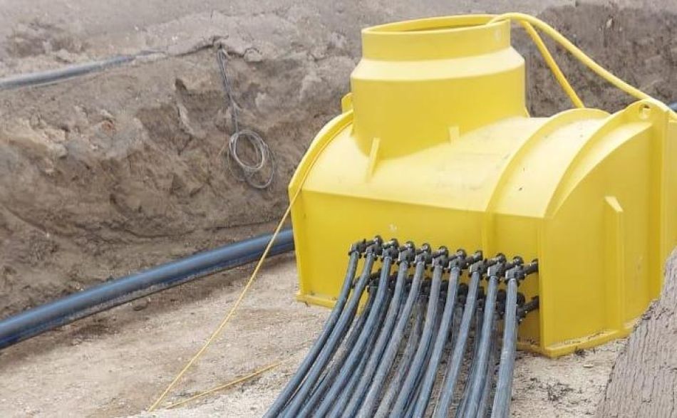 Ein leuchtend gelber unterirdischer Verteilerkasten in einem Erdgraben, der mit mehreren grauen Leitungen verbunden ist.