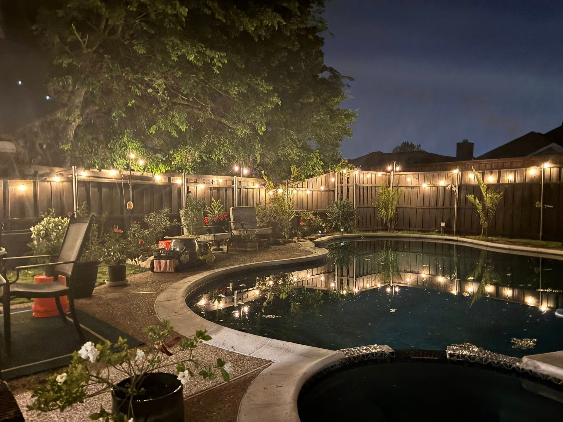 Lit backyard patio with string lights, pool, and chairs beside a tree at dusk