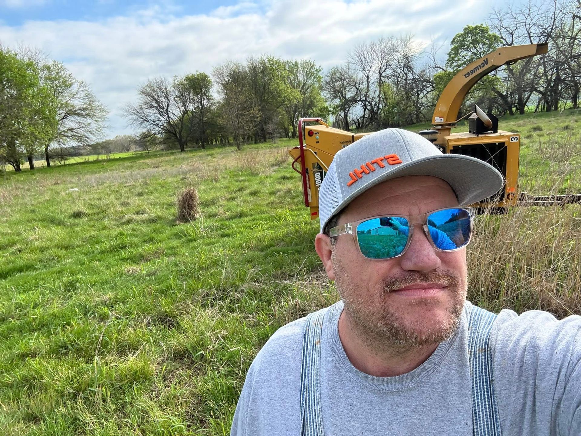Man wearing sunglasses and a cap in a grassy field with farm equipment in the background
