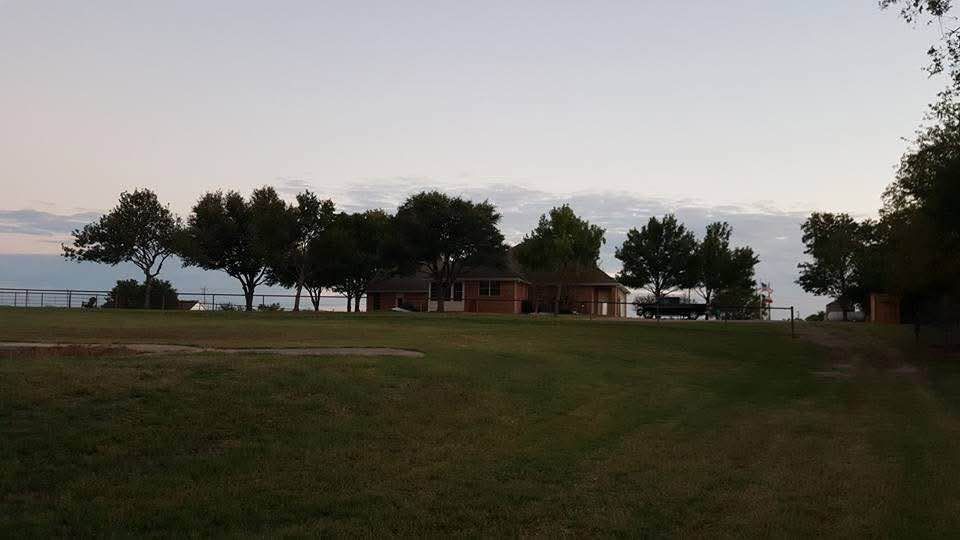 Grassy park with trees, a small building, and distant water under a cloudy sky