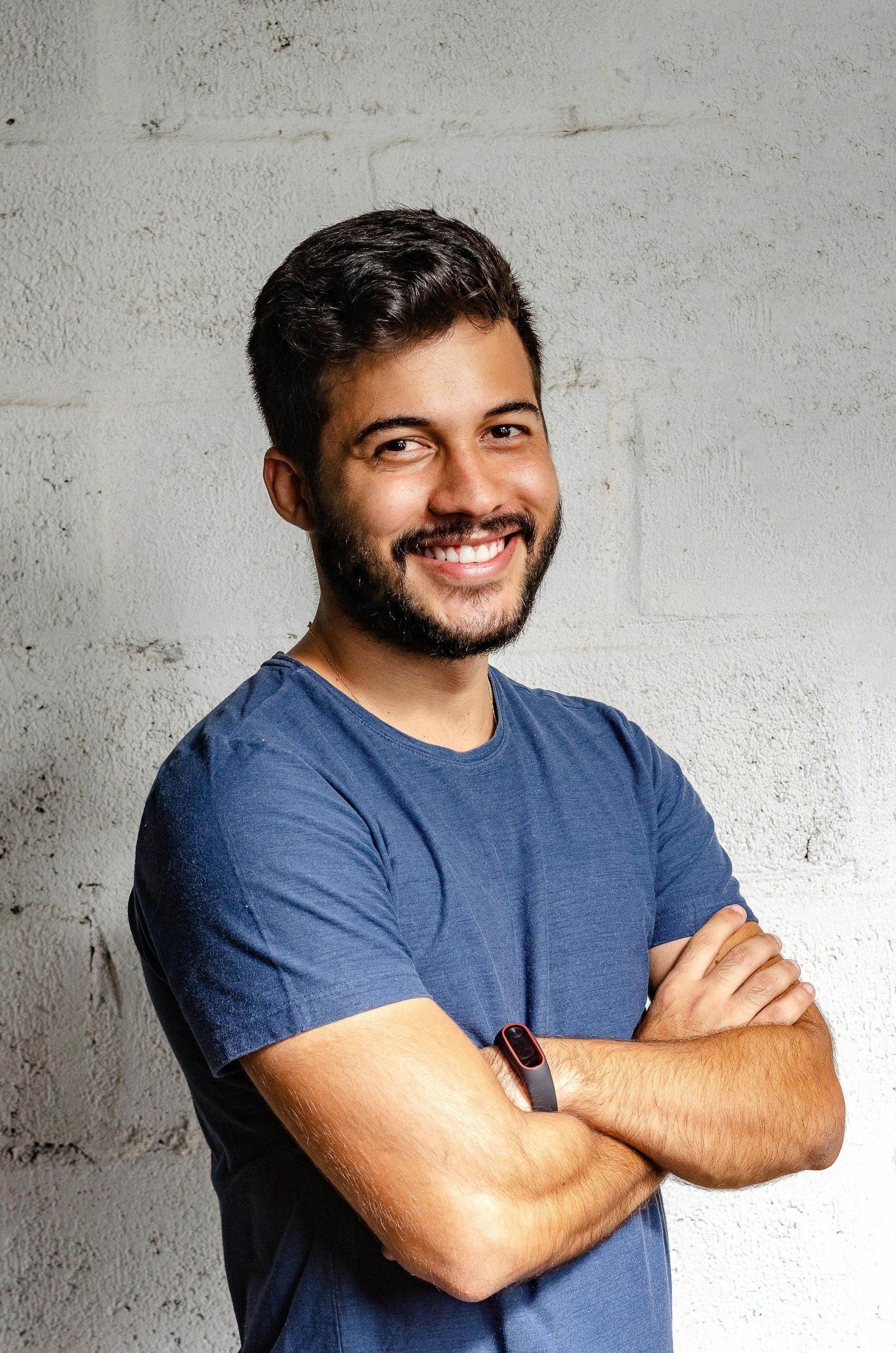 Un homme avec une barbe est debout, les bras croisés et souriant.