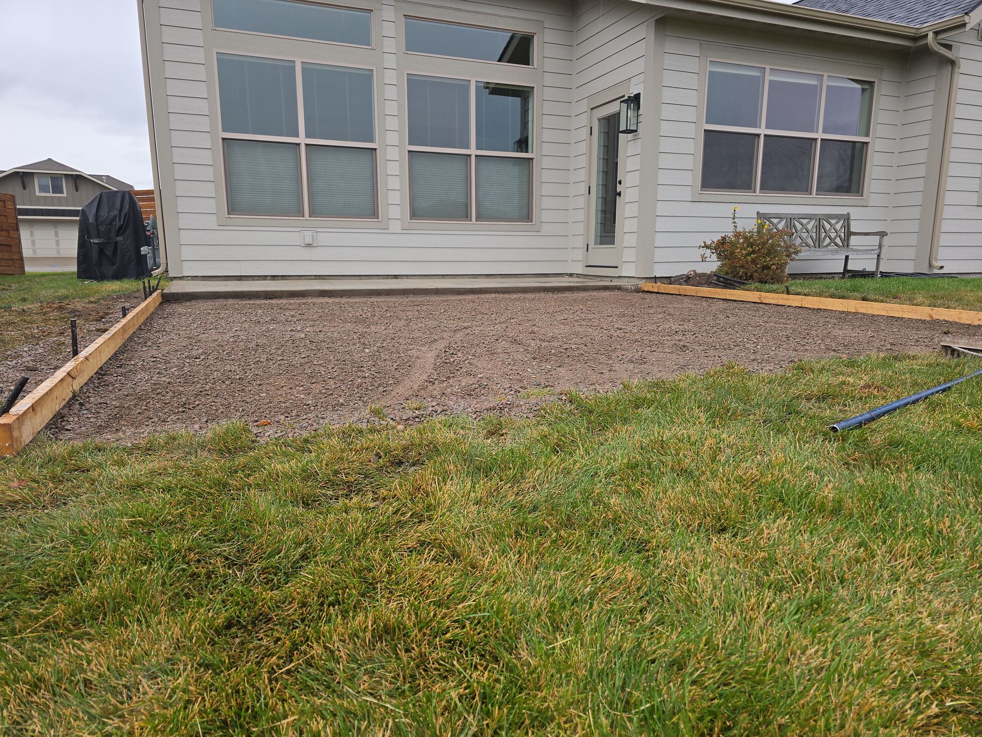 A gravel patio area framed by wooden forms, situated in the backyard of a house with beige siding and large windows.