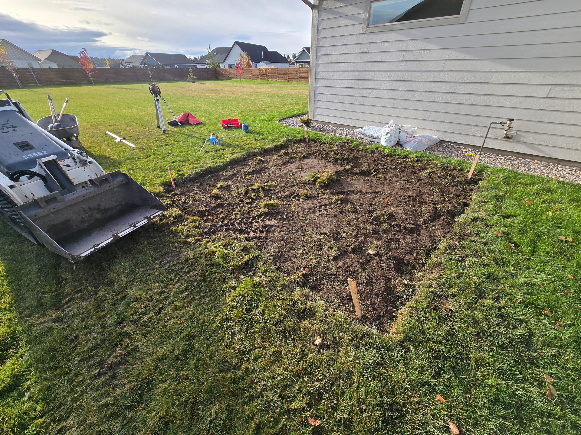 A skid steer loader sits near a square, dirt-excavated section in a grassy backyard beside a house.
