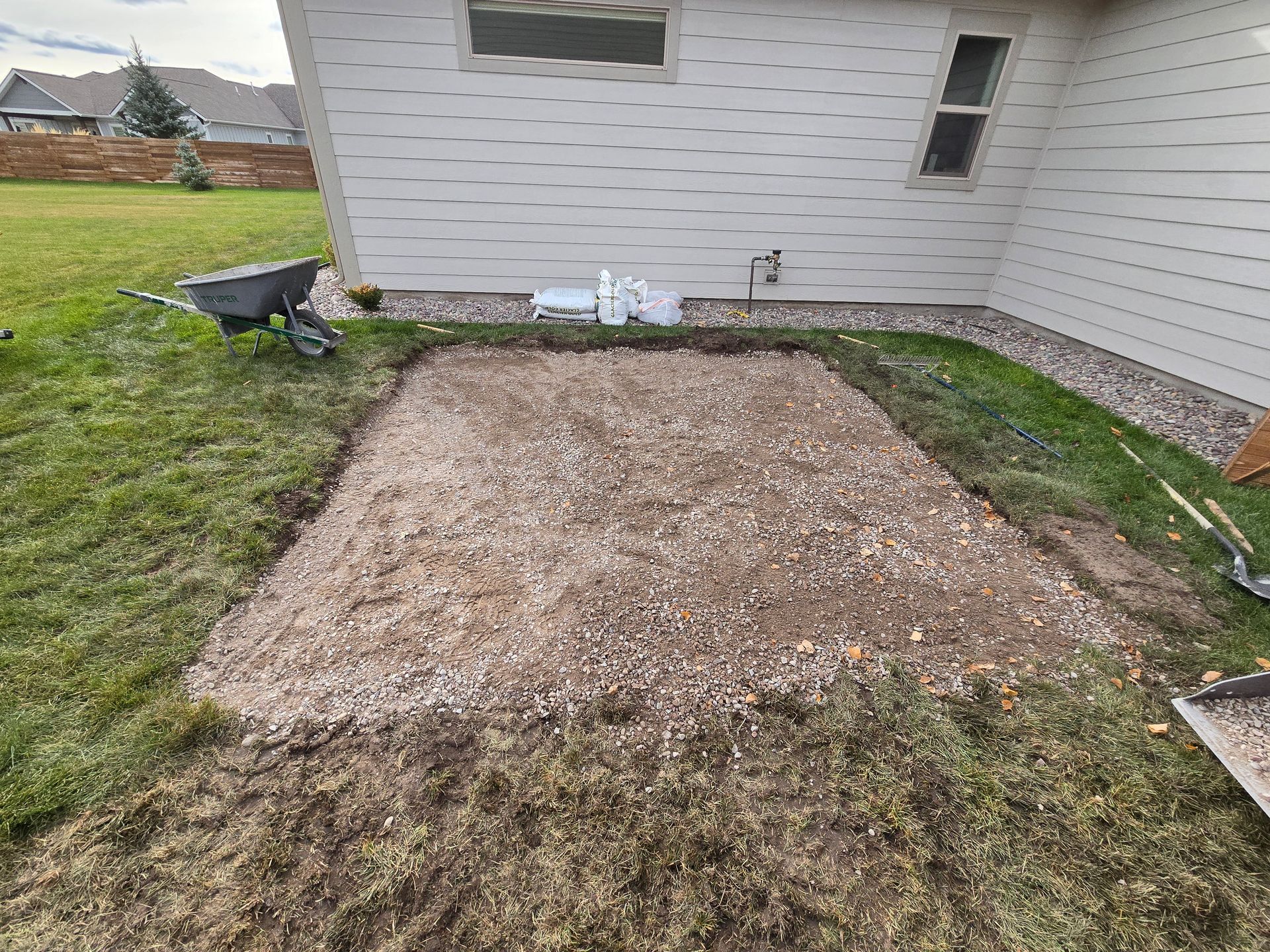 A rectangular gravel area prepared for construction next to a beige house, with a wheelbarrow nearby on the grass.