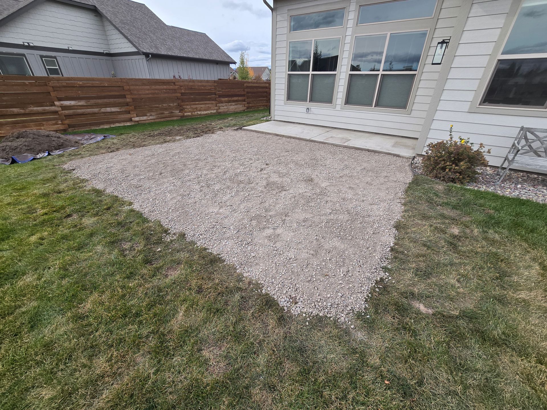 A rectangular gravel patio area sits in a backyard next to a house with large windows and a wooden fence in the background.