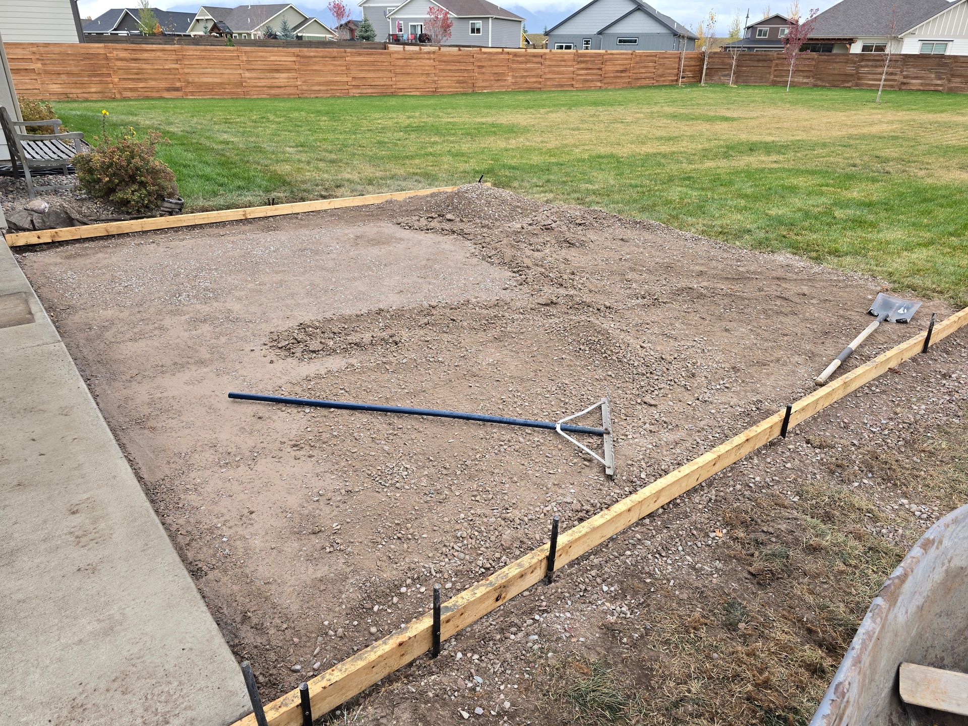 A dirt yard area prepared for construction with wooden forms set for a concrete pad and a rake lying on the ground.