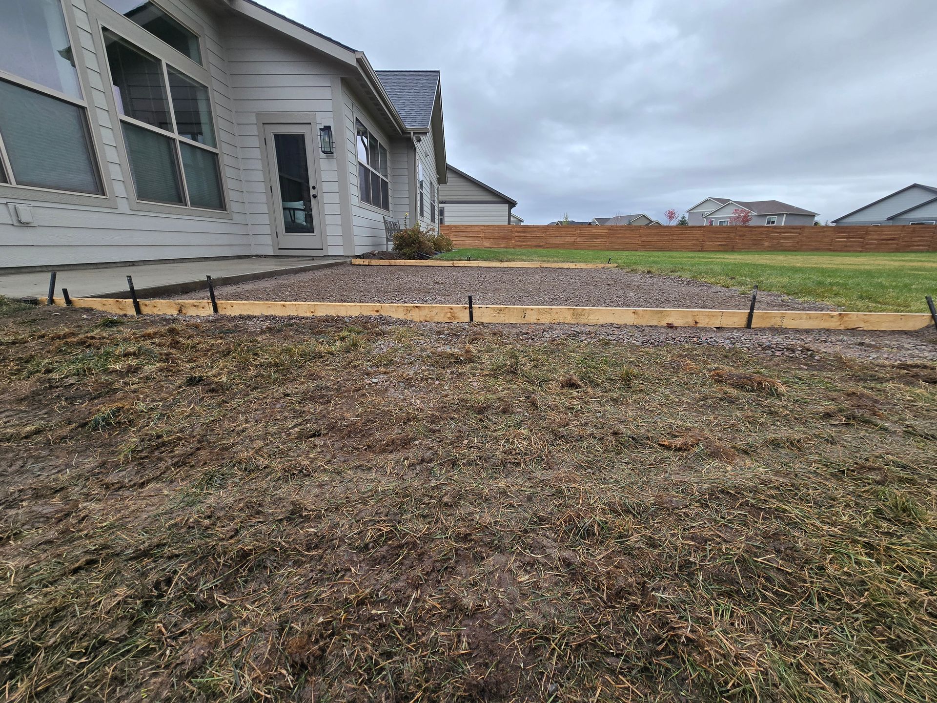 A gravel-filled rectangular area prepared with wooden forms for a patio extension next to a house under a cloudy sky.
