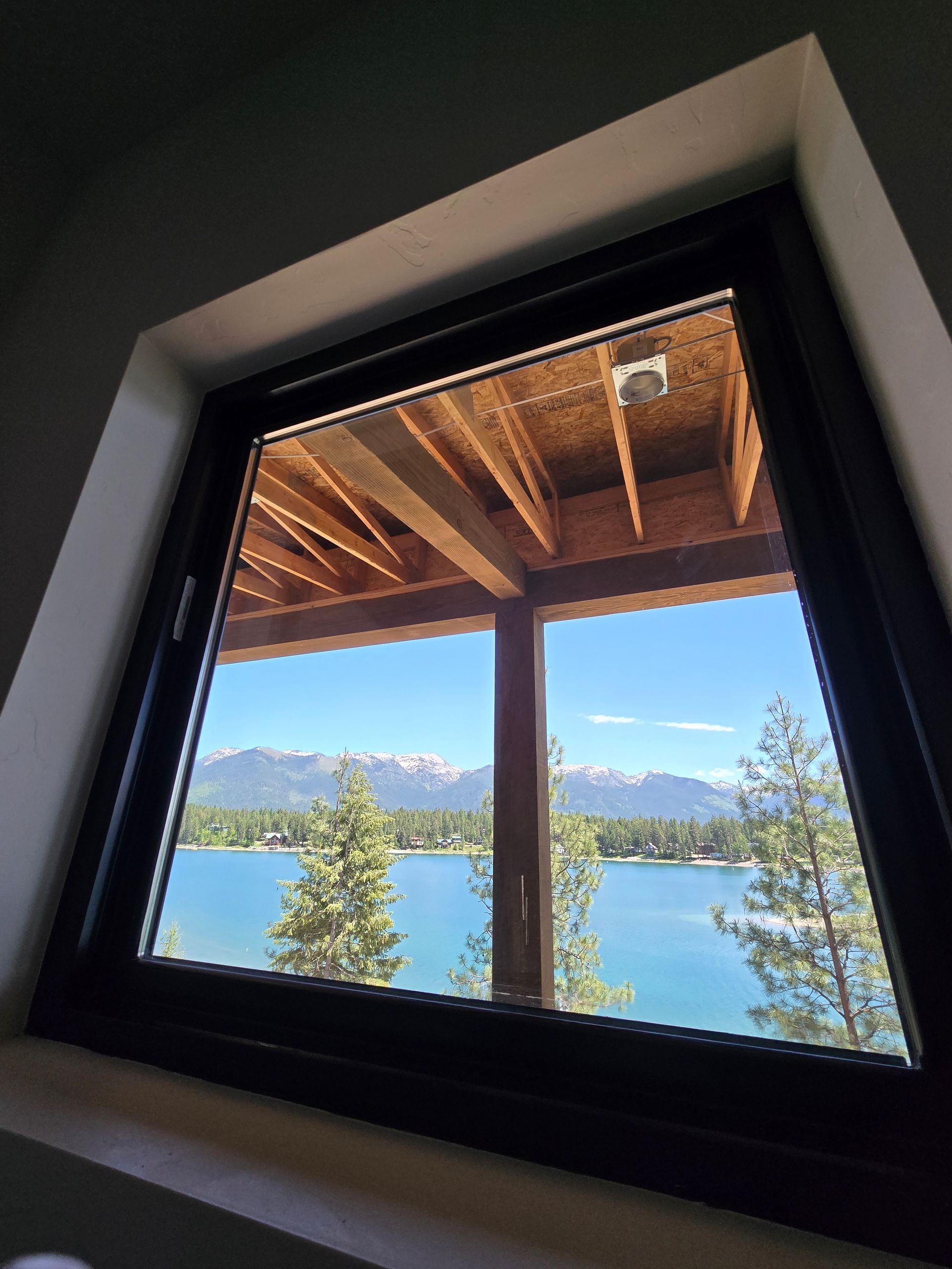 A view through a window showing a lake, pine trees, and snow-capped mountains under a bright blue sky.