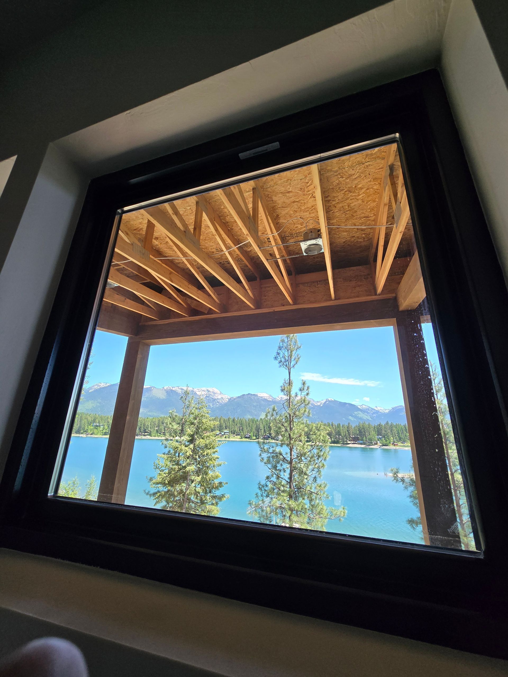 A view from inside through a window showing a wooden porch ceiling, a blue lake, pine trees, and distant snow-capped peaks.