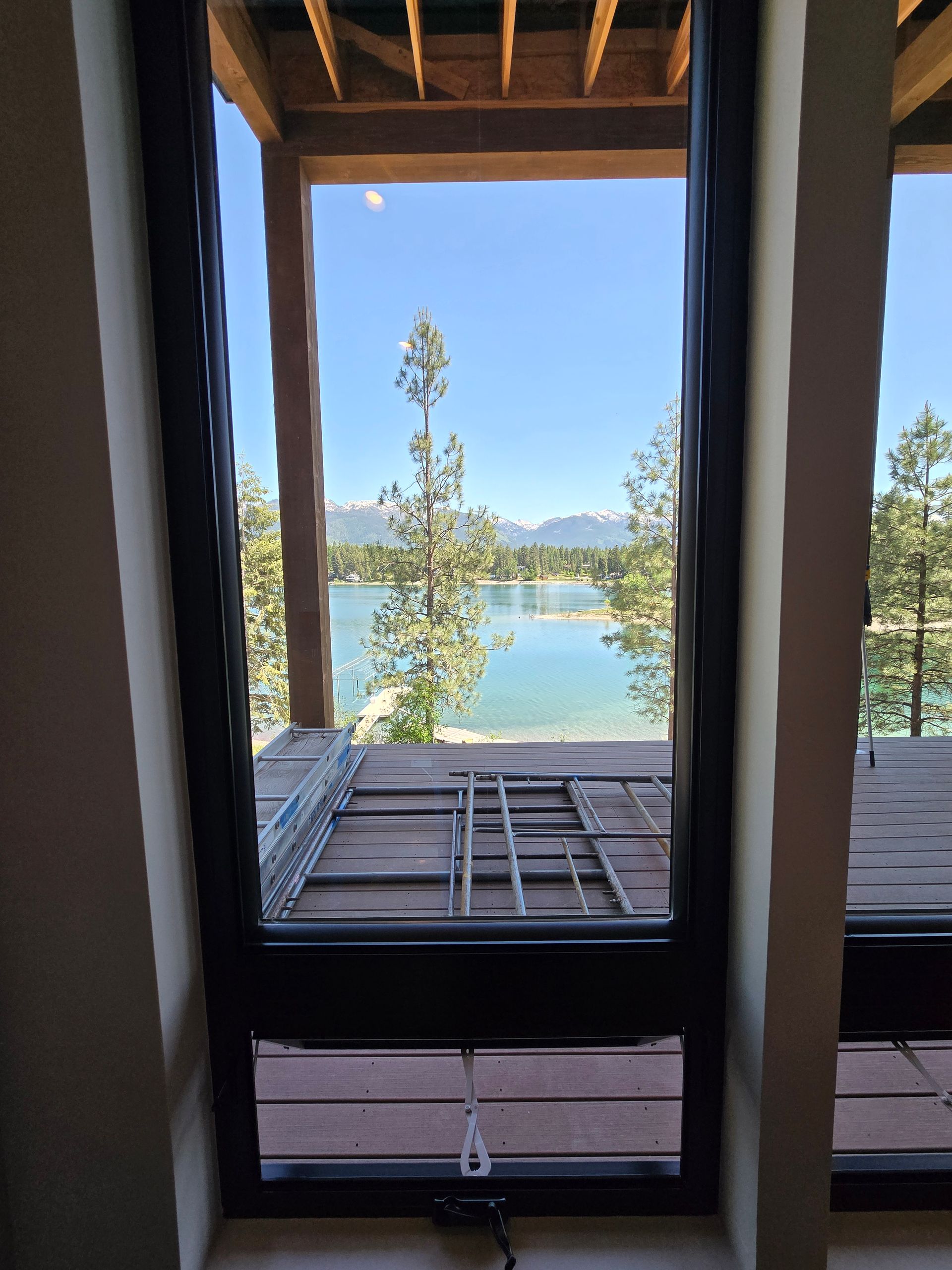 A tall, dark-framed window overlooks a wooden deck, a calm lake, trees, and distant mountains under a clear blue sky.