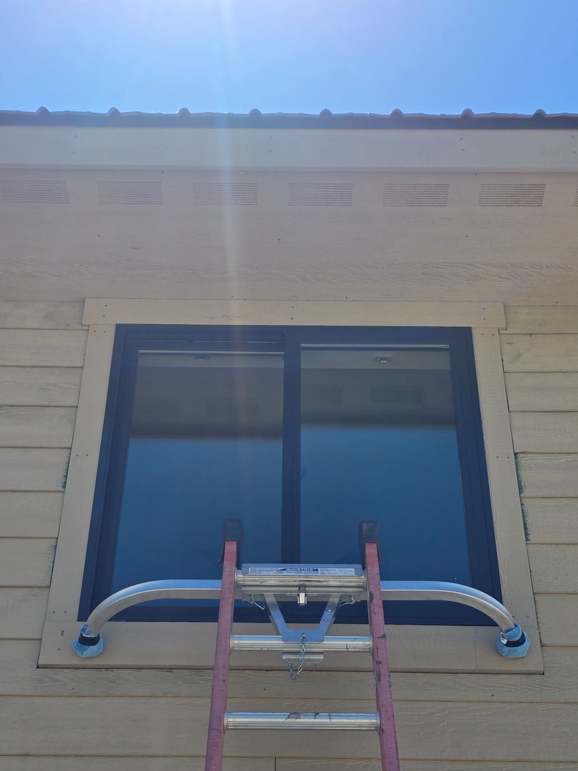A window stabilizer bar attached to a ladder, positioned against a house window under a sunny sky.