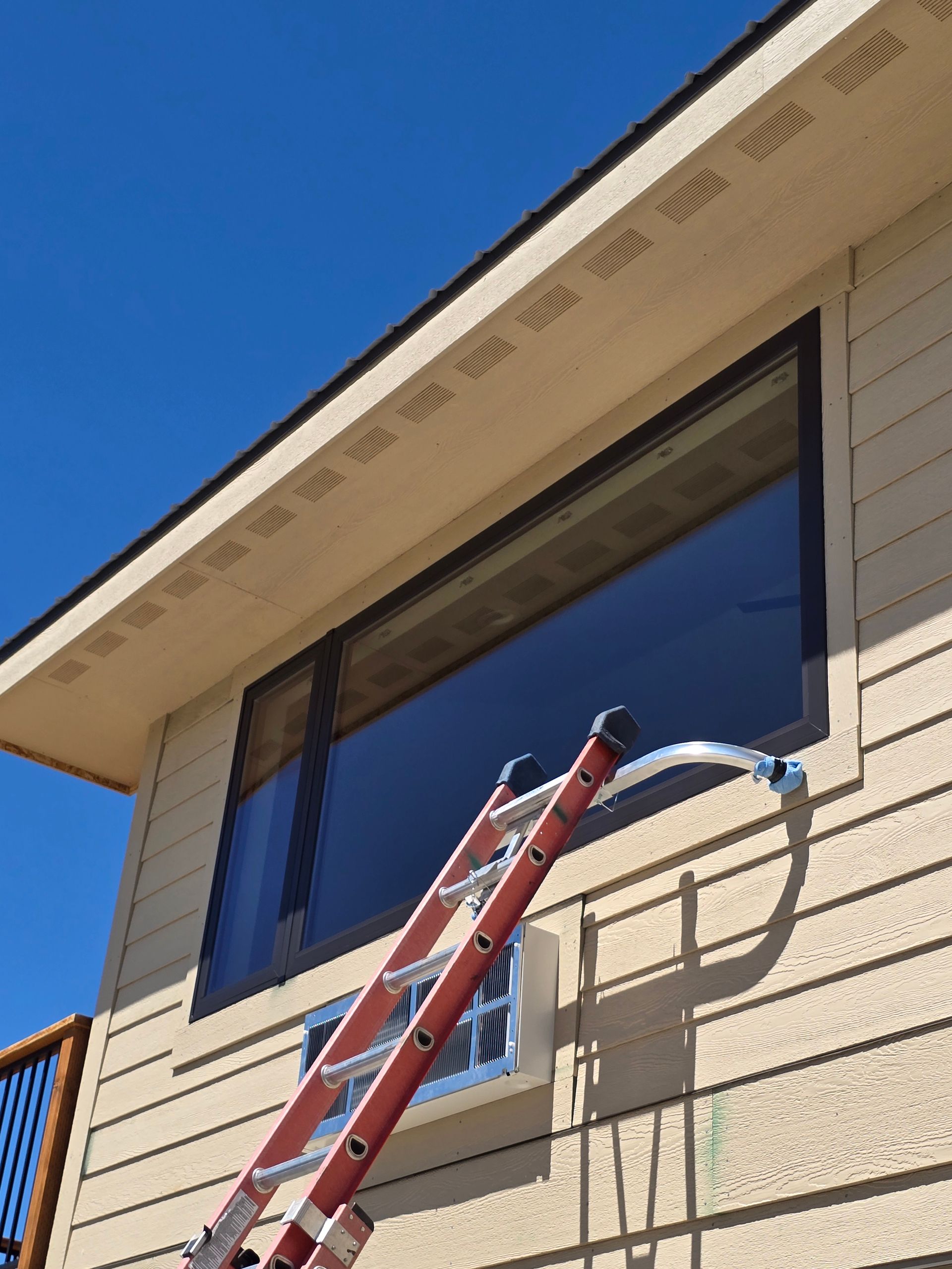 An extension ladder leans against the side of a tan house, positioned beneath a large window on a sunny day.