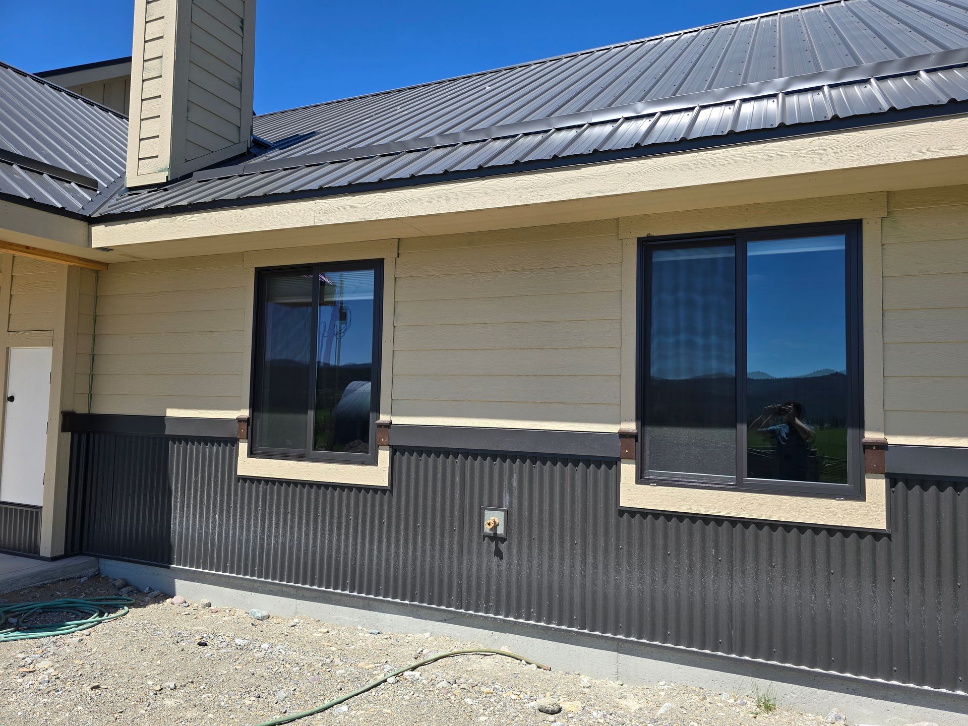 A residential building exterior with tan horizontal siding, dark corrugated lower paneling, and dark metal roof.