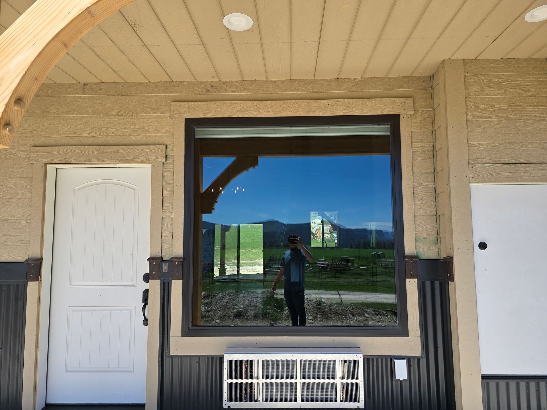 A building exterior features a white door, a large window reflecting a landscape, and an air conditioner below the window.