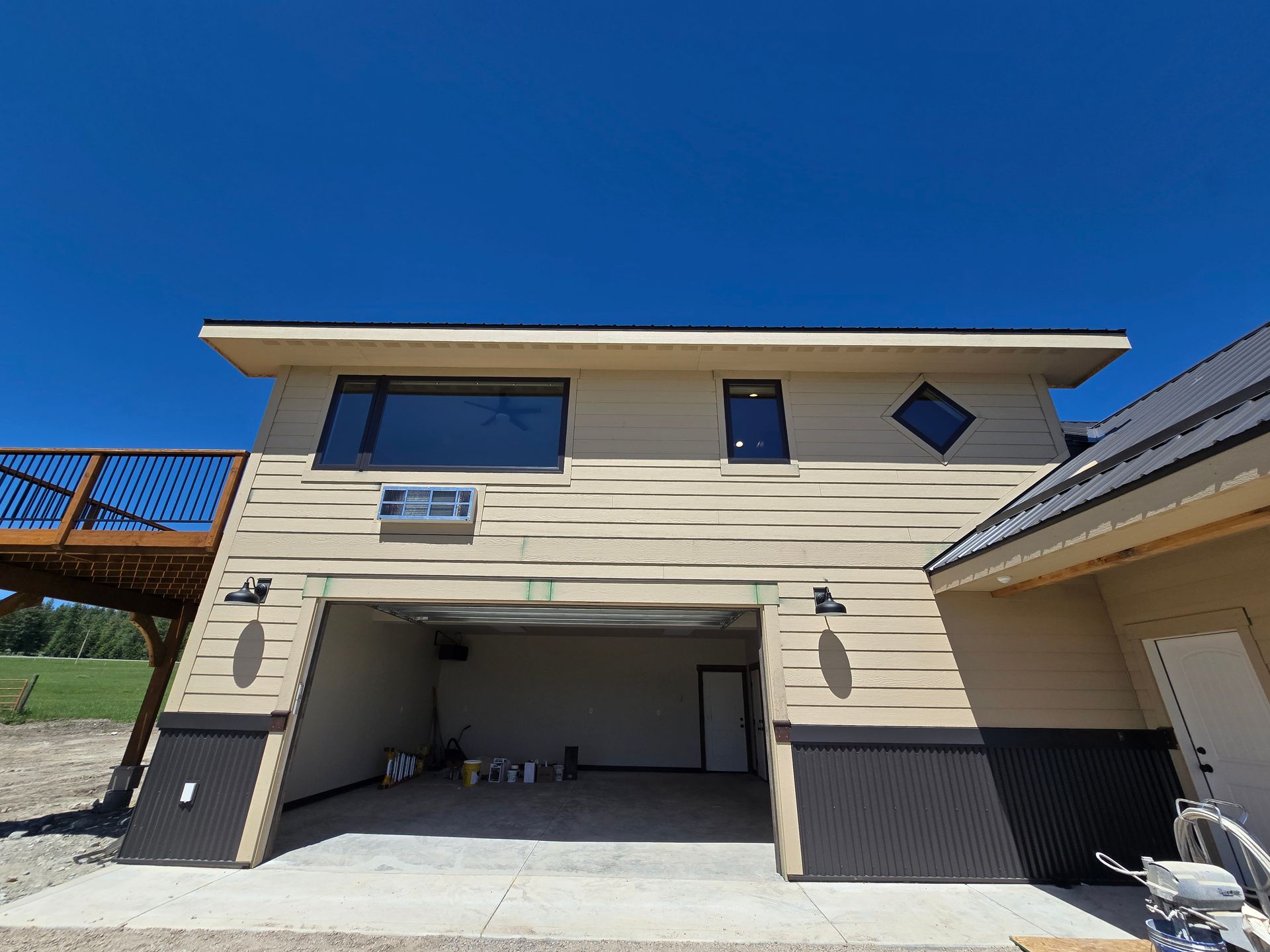 A tan two-story building featuring a large open garage bay, a deck, multiple windows, and a dark metal lower exterior.