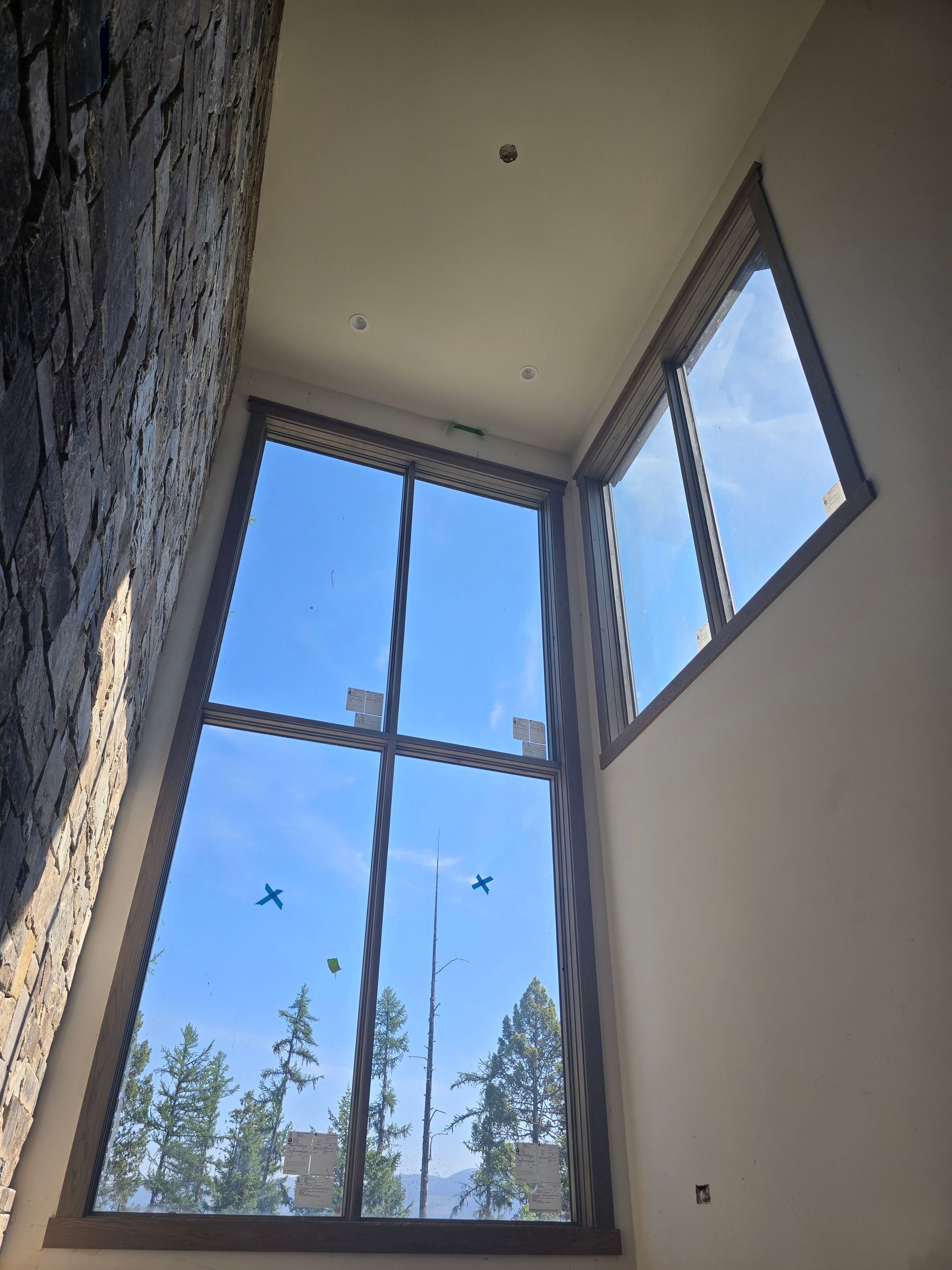 A tall corner window with decorative framing overlooks trees and a blue sky, next to a textured stone wall.