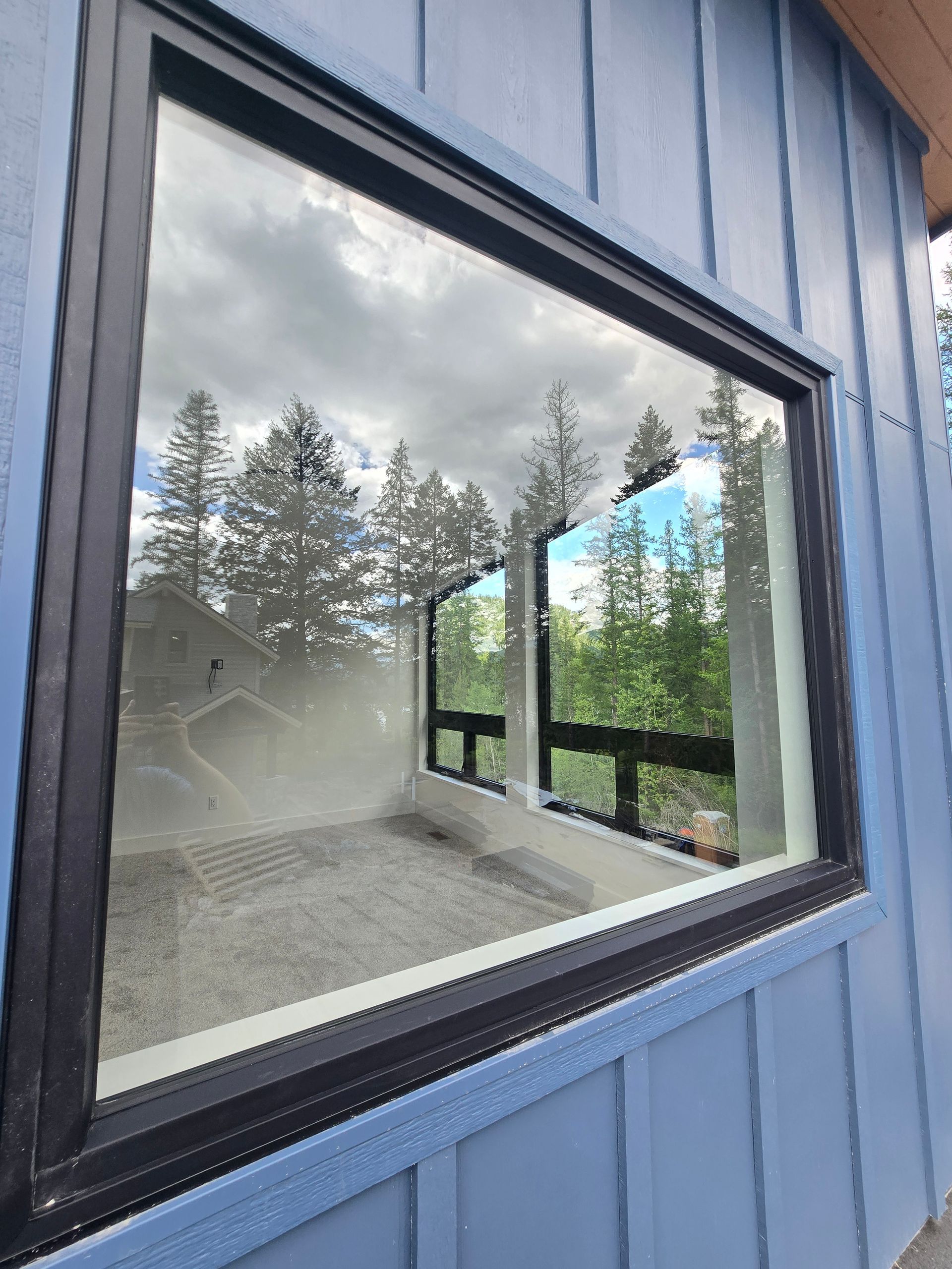 A large square window with a black frame reflecting trees and a blue sky, set into blue vertical siding.