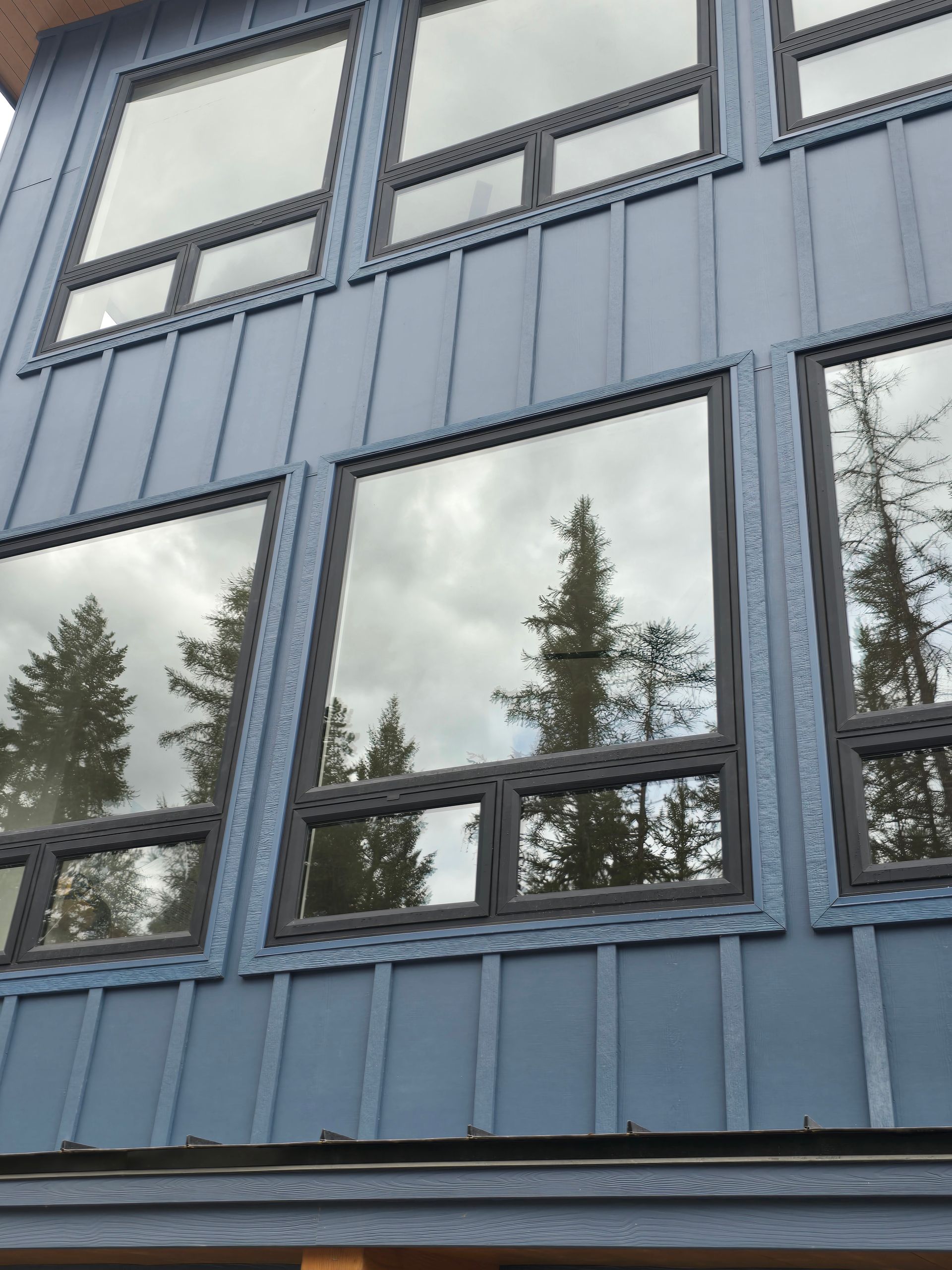 A low-angle view of a blue, board-and-batten-style exterior wall featuring large, reflective windows showing pine trees.
