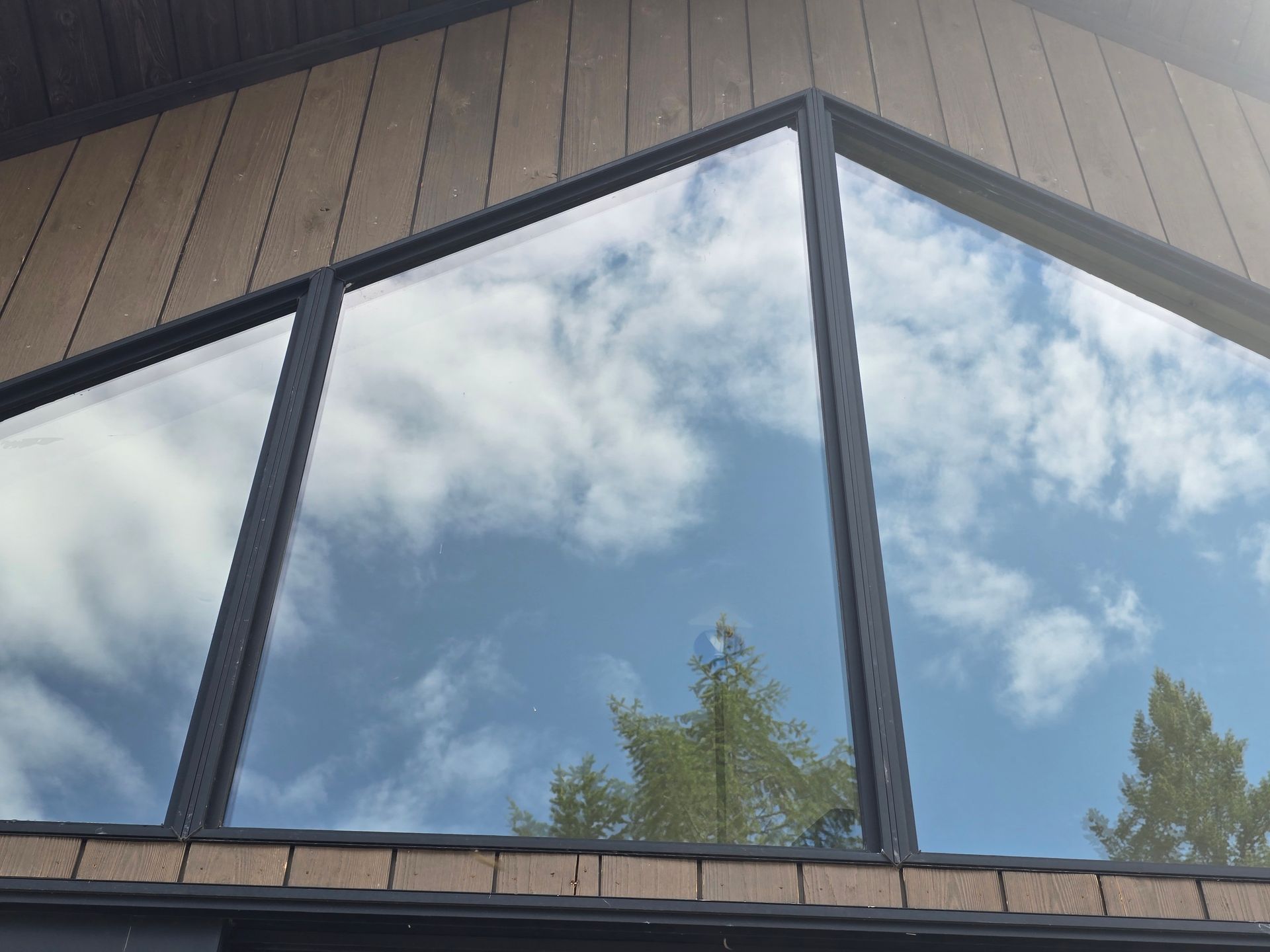 An angular, modern window set into a wooden-paneled wall, reflecting the blue sky, white clouds, and nearby trees.
